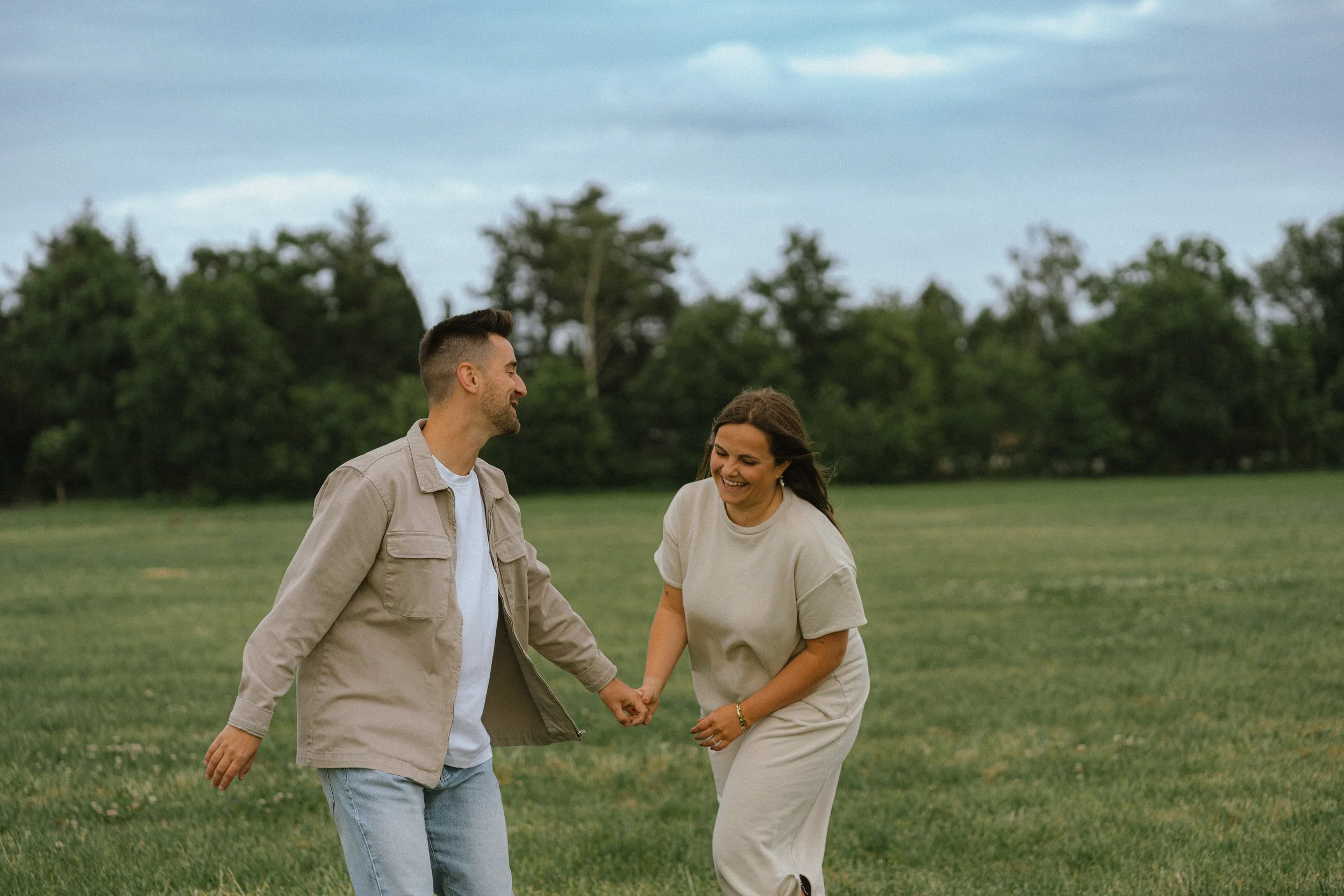 Man en vrouw lopen samen hand in hand op een grasveld, lachen en lijken te genieten van de dag, met bomen en een bewolkte lucht op de achtergrond.