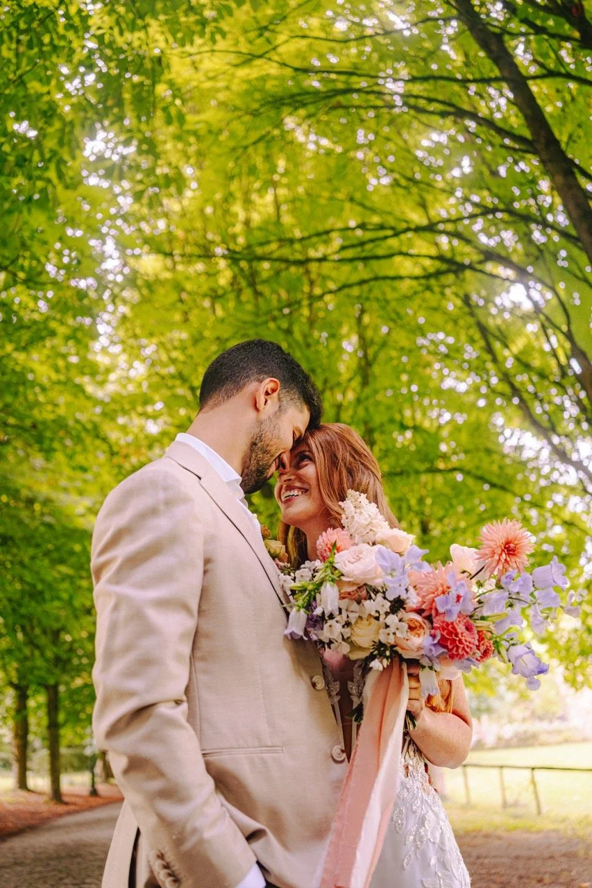 Koppel op een park, geïnterviewd op hun bruiloft, omringd door groene bomen, zij met een boeket bloemen, hij in een beige pak, glimlachend naar elkaar.