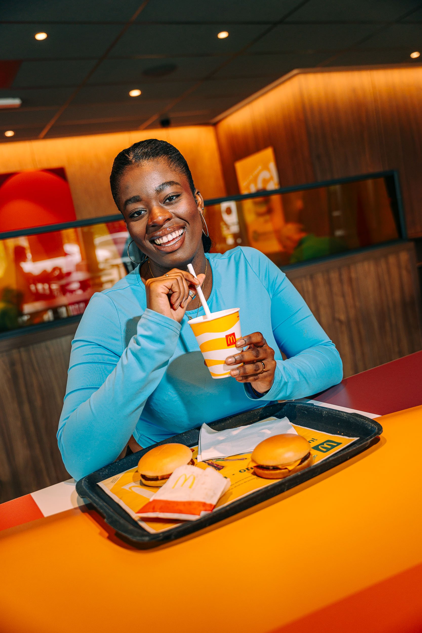 Vrouw glimlachend in een McDonald's restaurant, drinkend uit een papieren beker met een rietje, met twee cheeseburgers en een maaltijddoos op een dienblad voor haar.