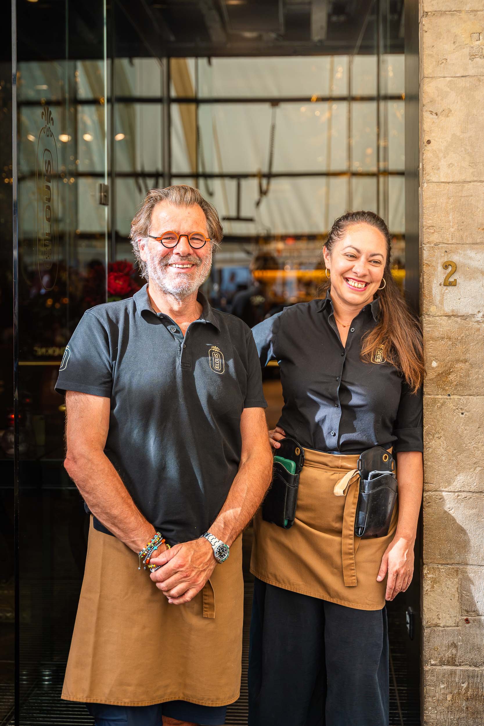 Twee mensen, een man en een vrouw, staan voor een glazen deur met het nummer 2, beiden dragen zwart uniform met een logo en glimlachen.
