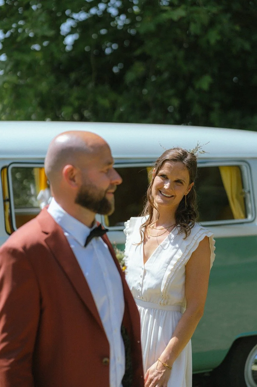 Een man en een vrouw glimlachen en staan buiten voor een camper, waarschijnlijk tijdens een bruiloft of feest. De vrouw draagt een witte jurk en de man een bruiloftsjas.
