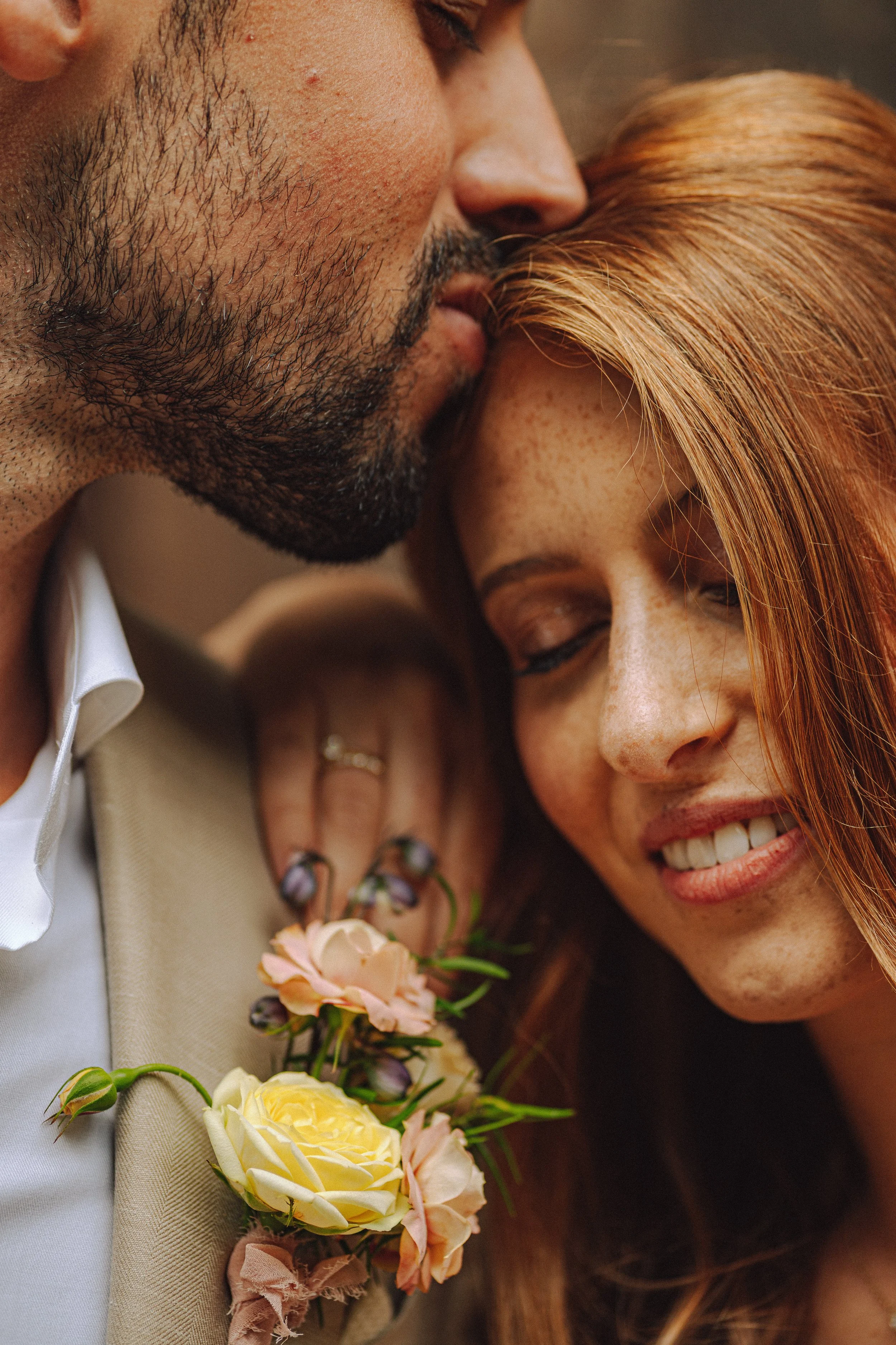 Close-up van een jong stel dat dicht bij elkaar staat, lachen en een kus uitwisselt. De vrouw heeft rood haar en een bloemenbroche op haar jurk, de man heeft een licht beige pak en een bloemencorsage.