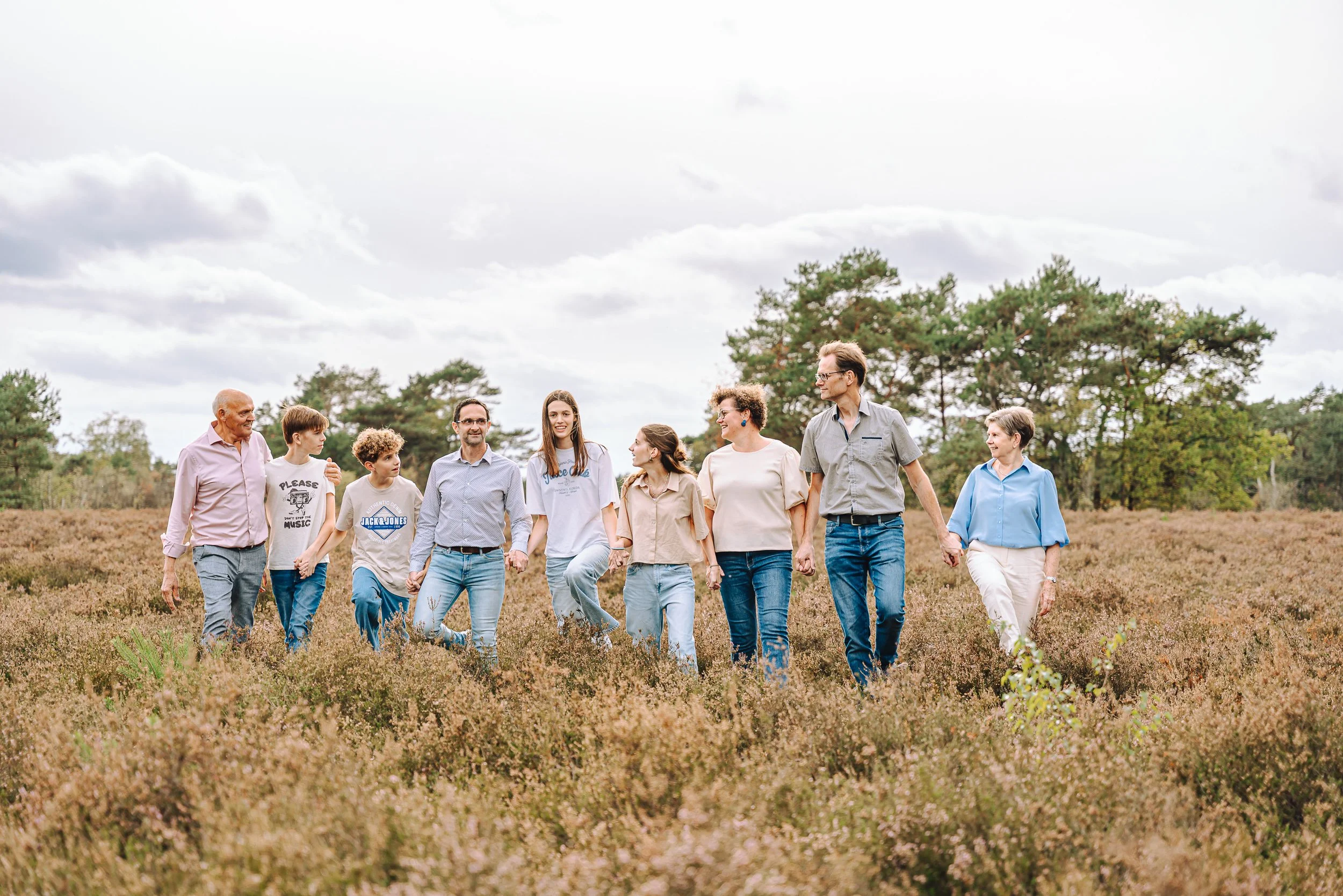 Een groep mensen wandelt samen door een veld met lage planten, onder een bewolkte hemel, in een bosrijke omgeving.