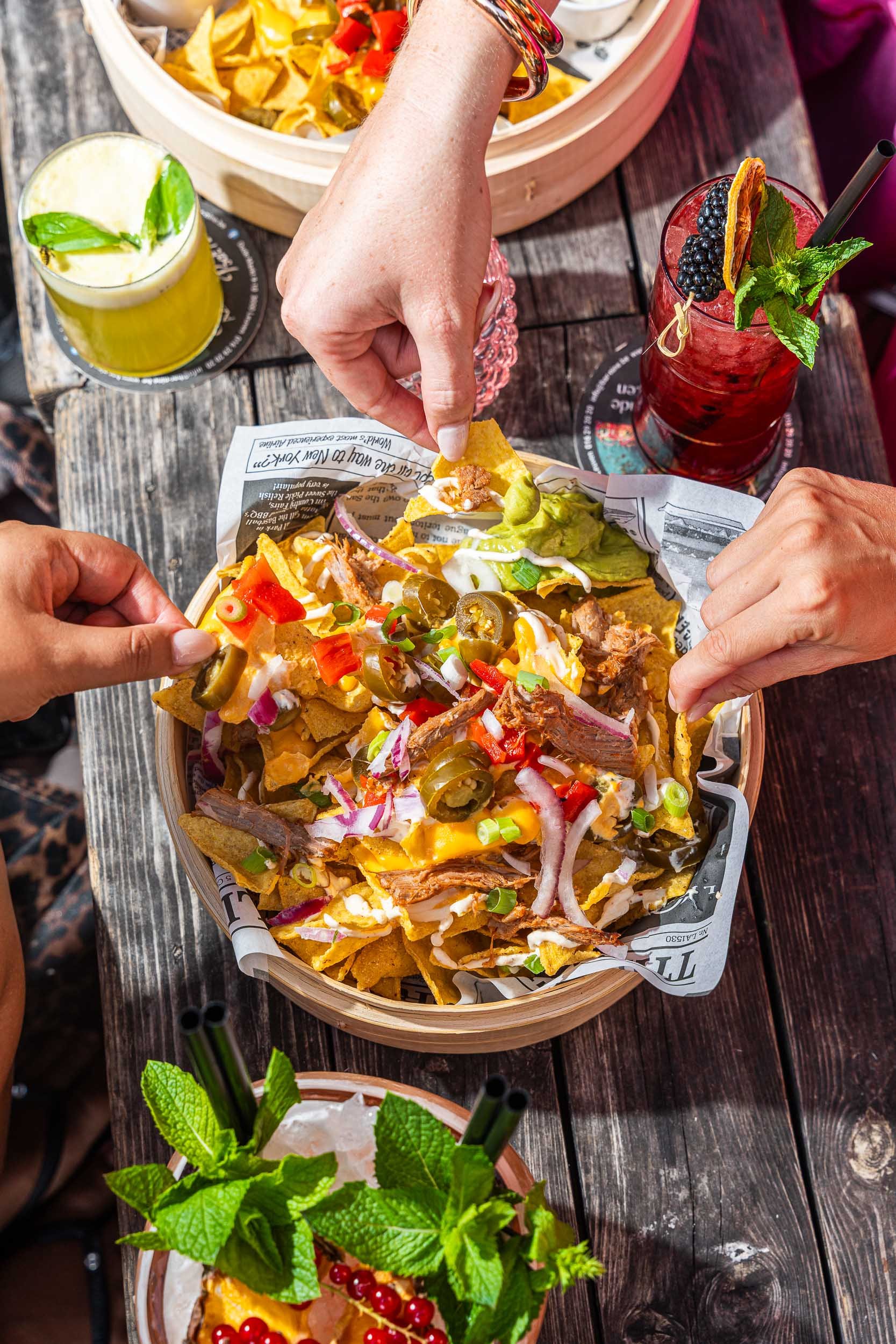 Verschillende handen die genieten van een bord met nachos bedekt met kaas, vlees, ui, paprika, olijven, en koriander, met drankjes op een houten tafel.