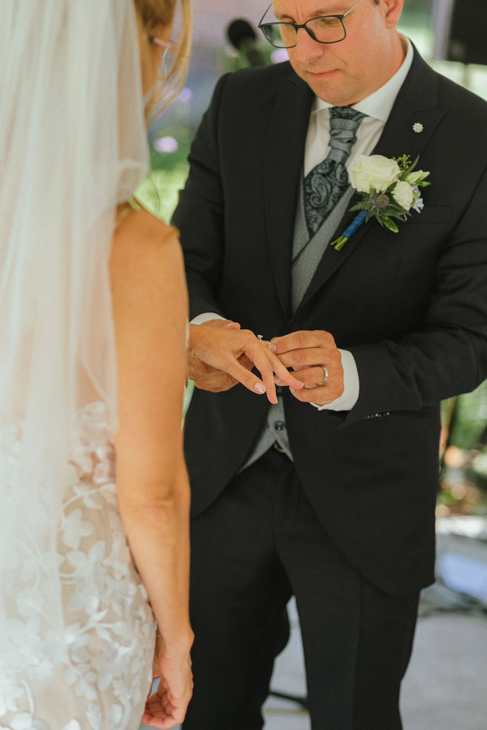 Een man en een vrouw tijdens een huwelijksceremonie, de man legt een ring om de vinger van de vrouw, de vrouw draagt een witte jurk met kant.