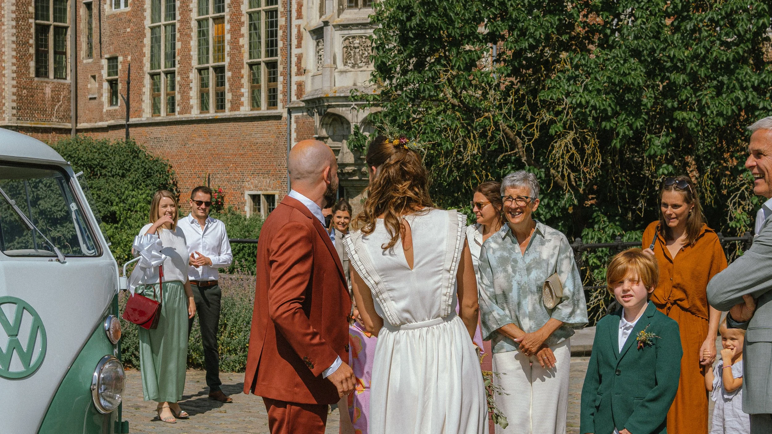 Groep mensen die samen lachen en praten buiten voor een historisch gebouw, met een witte en groene Volkswagen bus op de voorgrond.