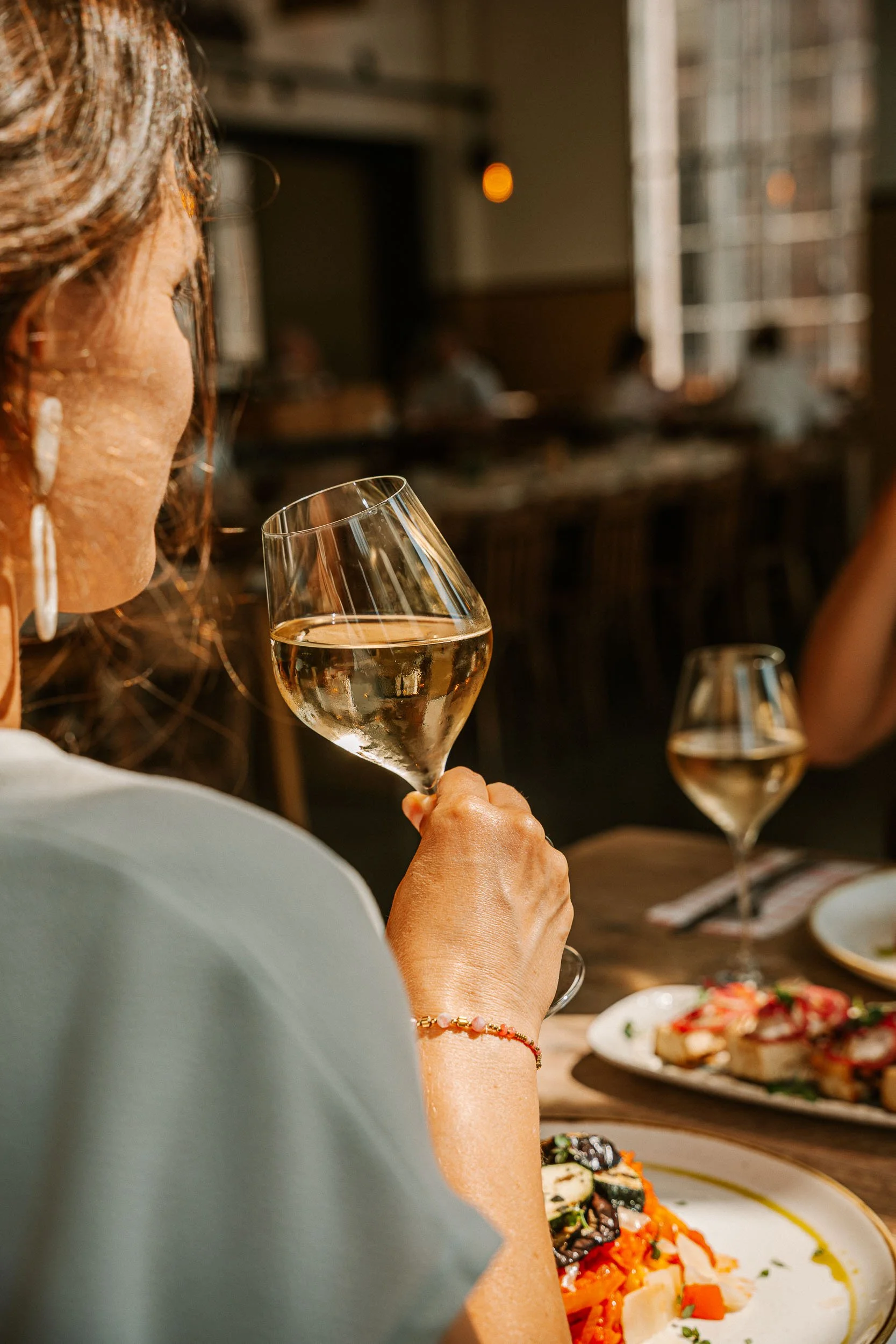Een persoon die in een restaurant of café wijn drinkt, met een bord met eten op de tafel voor hen, en anderen die aan tafel op de achtergrond zitten.