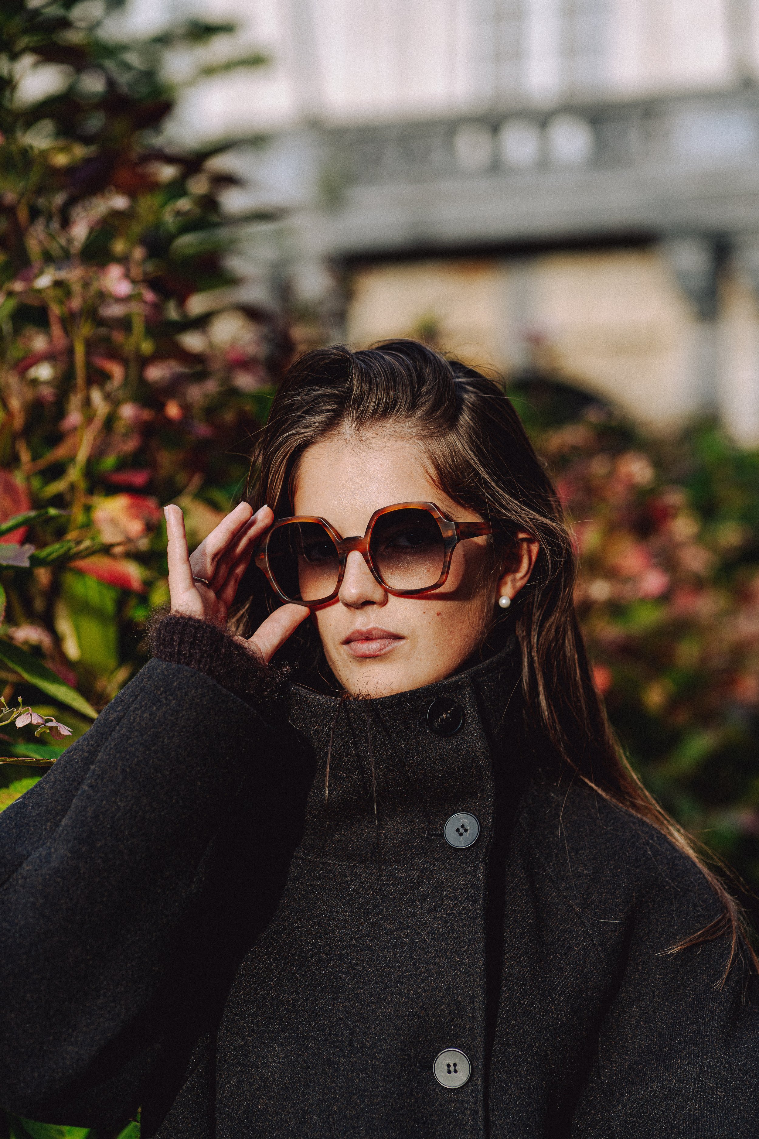 Een vrouw met bruin haar en grote ronde zonnebril die haar hand naar haar gezicht brengt, zittend in een tuin onder een overkapping met bladeren en bloemen op de achtergrond.