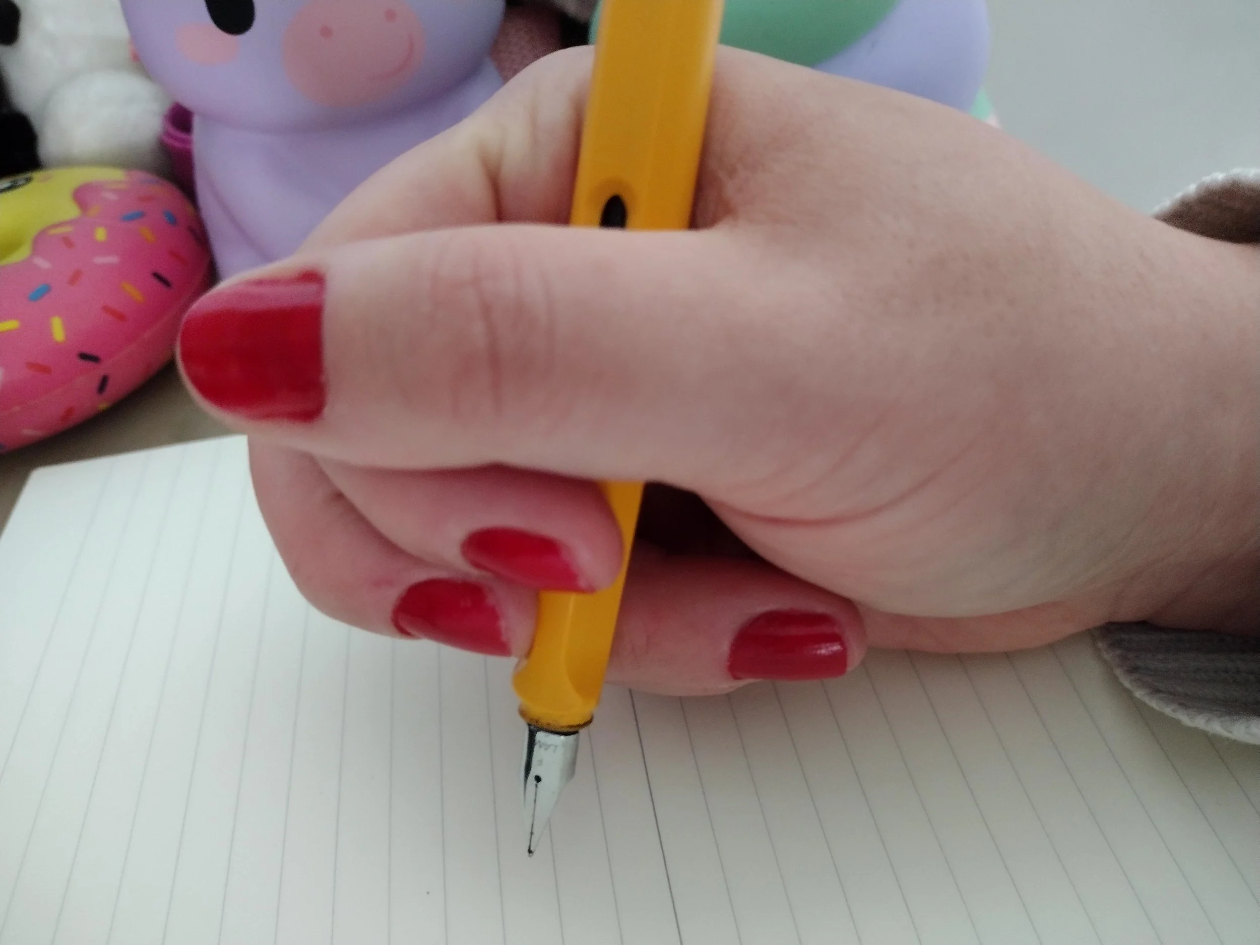 Kaye Outhwaite's hand gripping a yellow dip pen in a closed fist-style hold, pen angled diagonally across the palm with the nib pointing back toward the wrist, demonstrating an unconventional Dysgraphic pencil grip.
