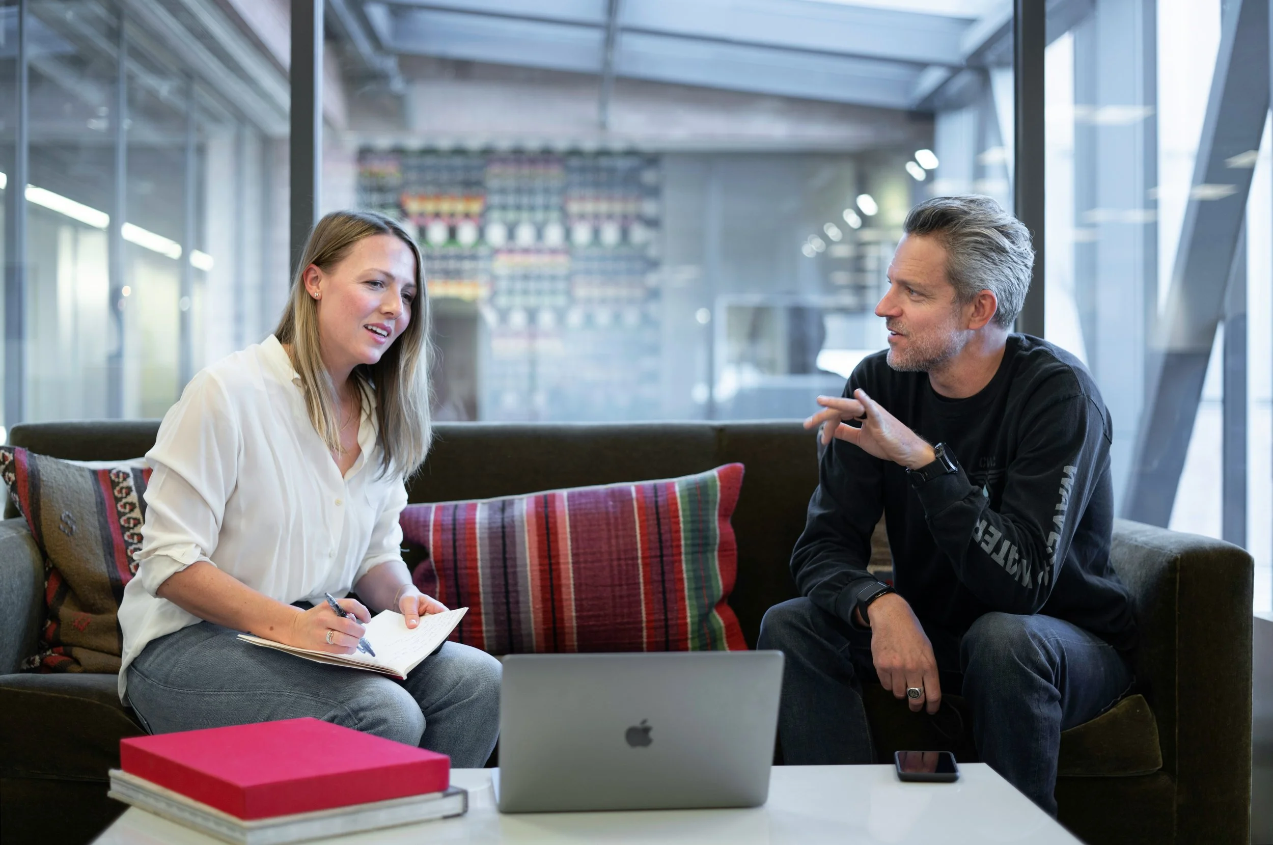 Two people, a woman and a man, engaged in a conversation while sitting on a sofa in a modern office space. The woman is taking notes in a notebook, and there is a laptop on the table in front of them, representing an sales process.