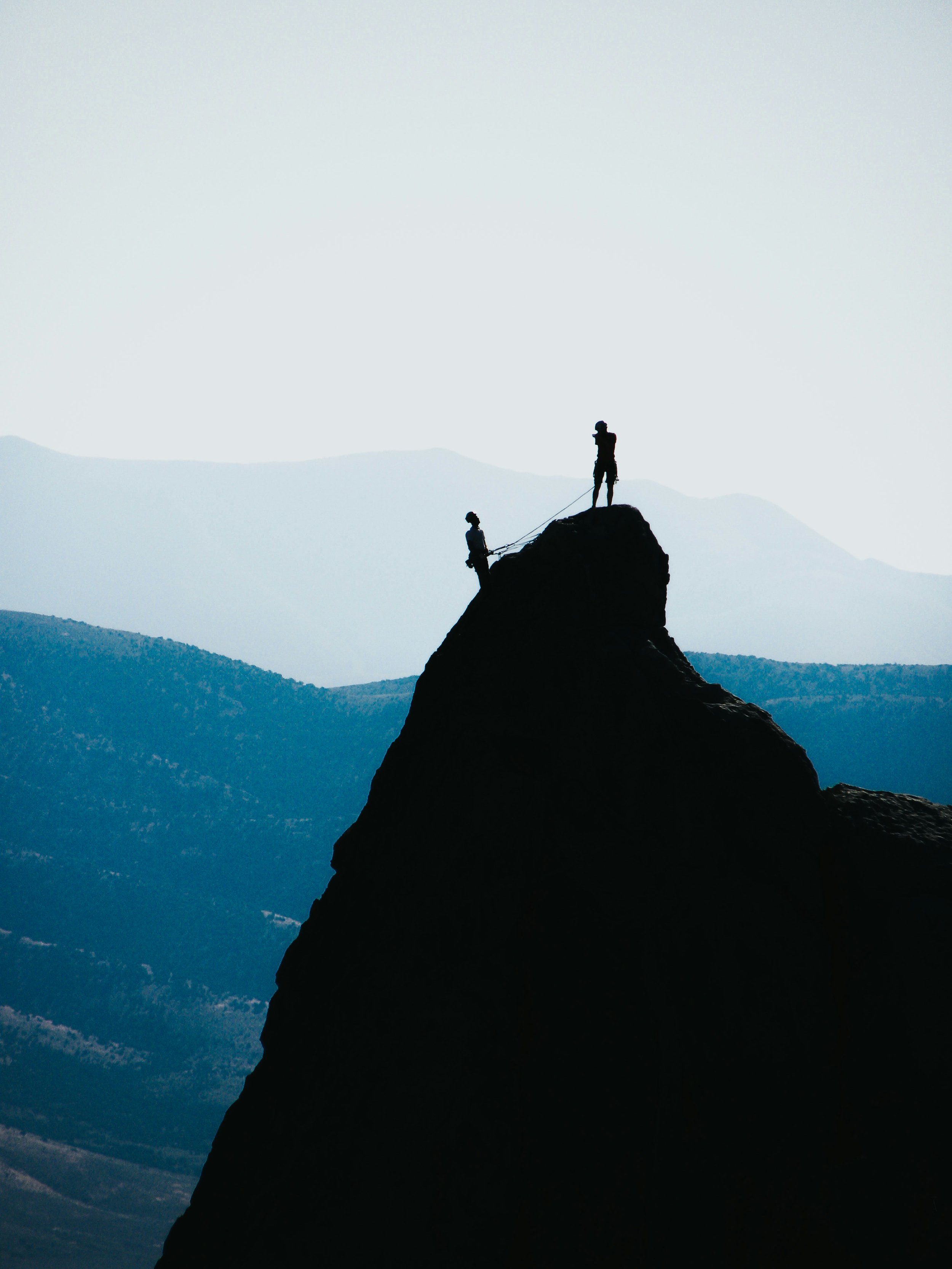 A silhouette of two rock climbers on a mountain summit with a mountain range in the background, representing the development of leadership in assisting others to reach their best potential