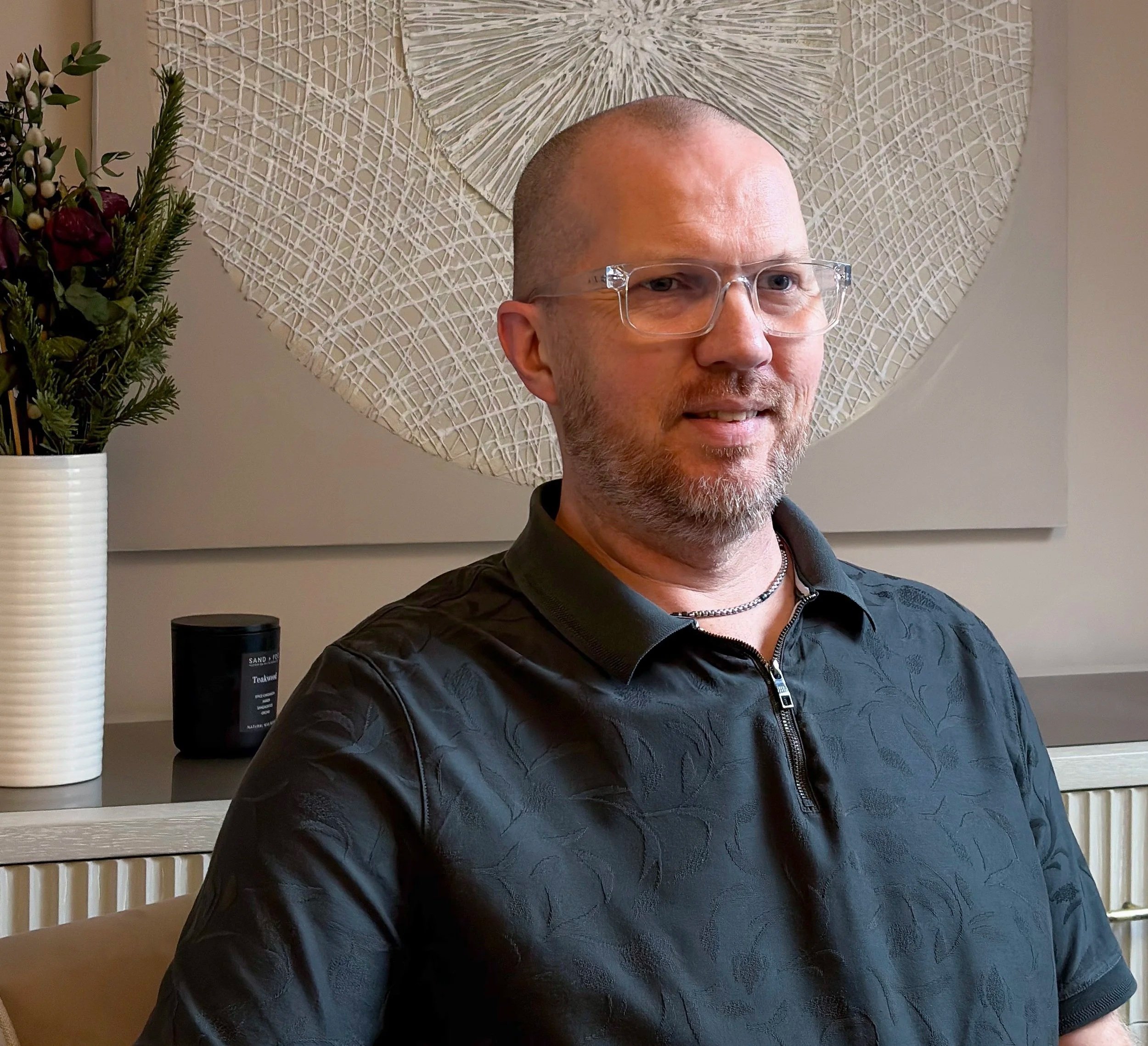 A man with a beard and glasses sitting indoors in front of a wall art and a vase with plants.