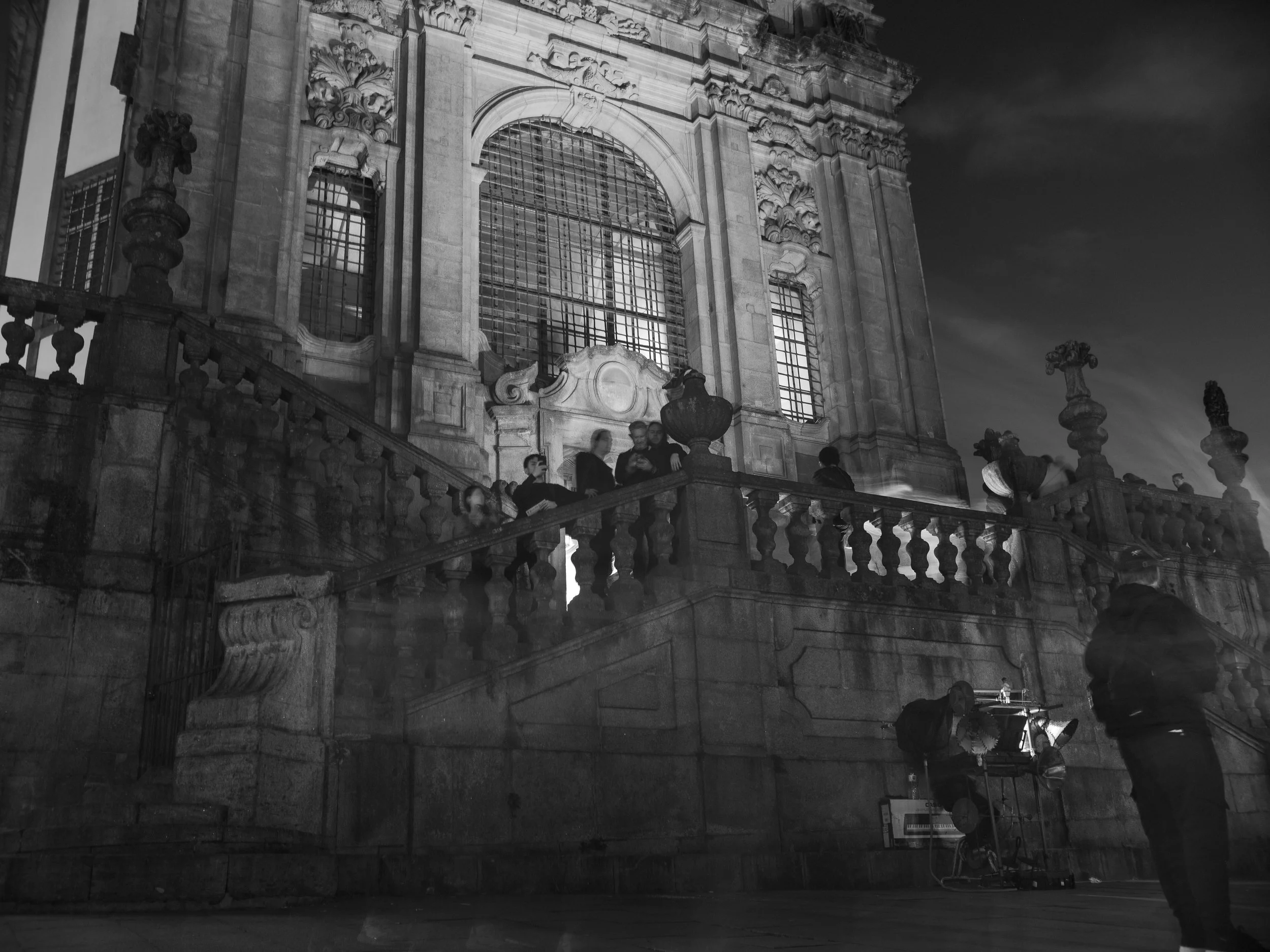 Nighttime scene outside a historic building with large ornate windows, stone staircase, and stone balustrade, where a group of people are gathered and a man operates a film camera on a tripod.