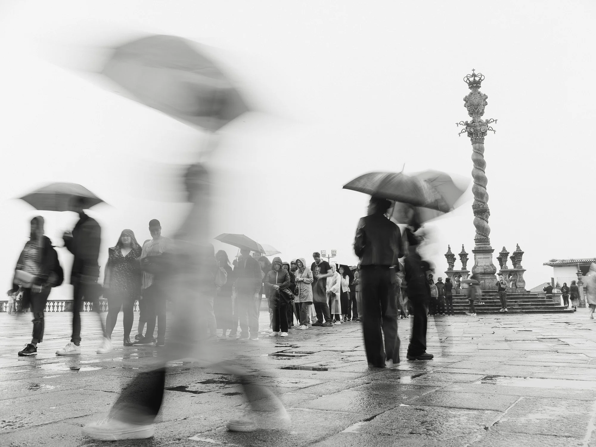 Black and white photo of people walking with umbrellas on a rainy day, some waiting in line, and a decorative lamppost in the background.