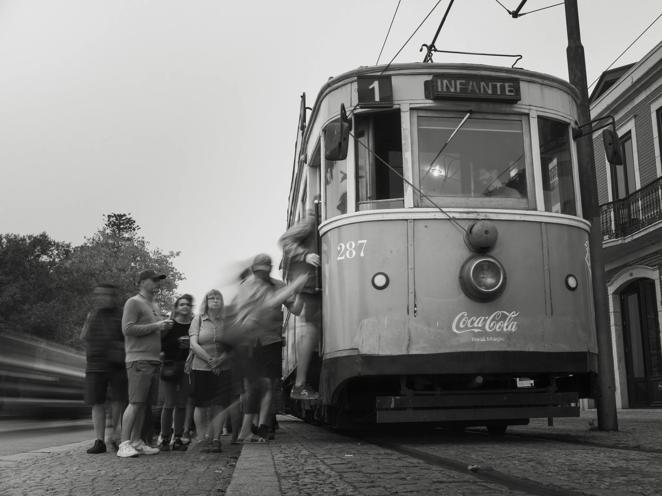A black and white photo of a vintage streetcar with the destination sign 'INFANTE' and Coca-Cola branding on the front, as people board and wait in line, with some in motion blur.