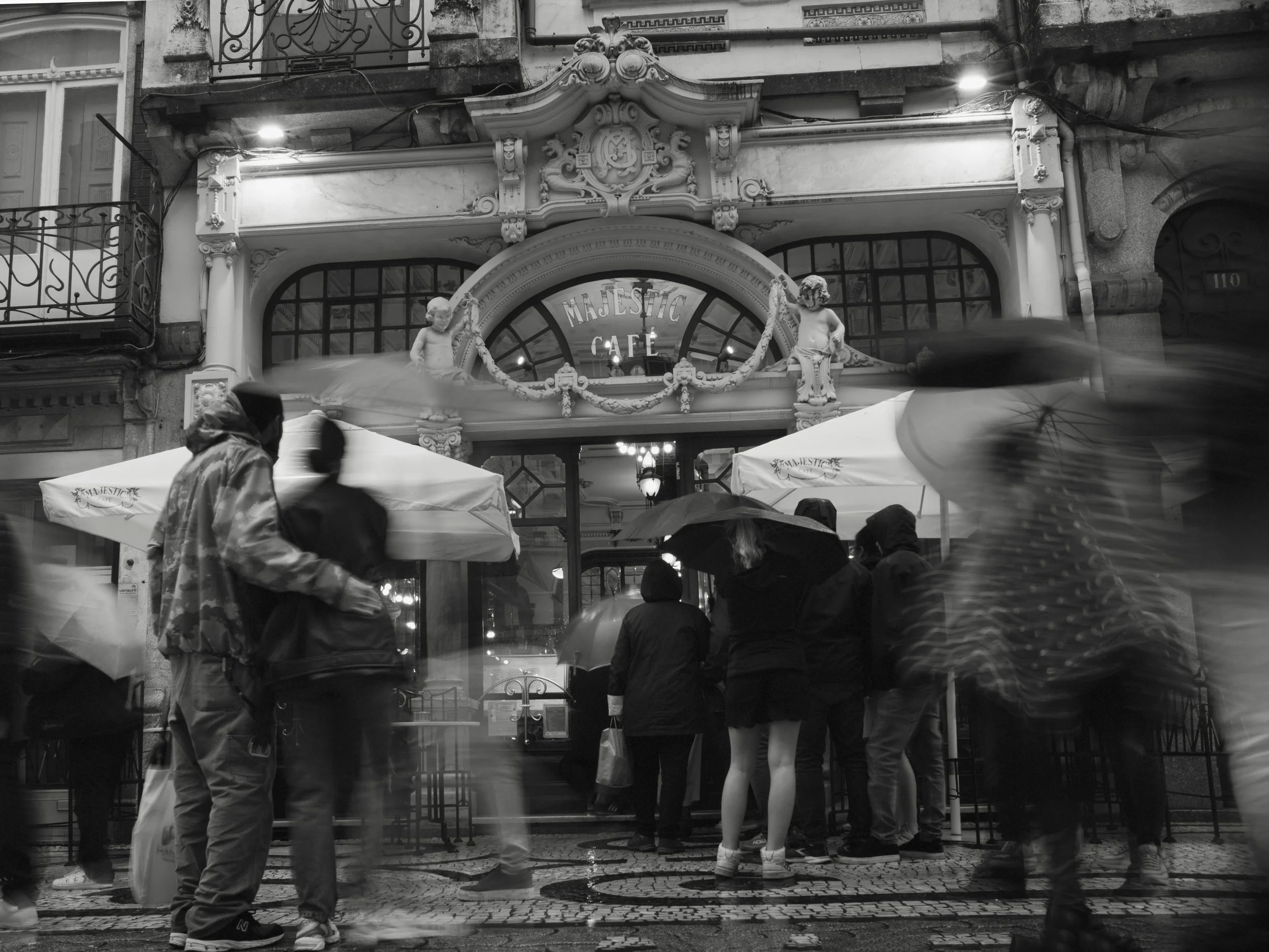 People with umbrellas wait outside a café on a rainy day, in black and white.