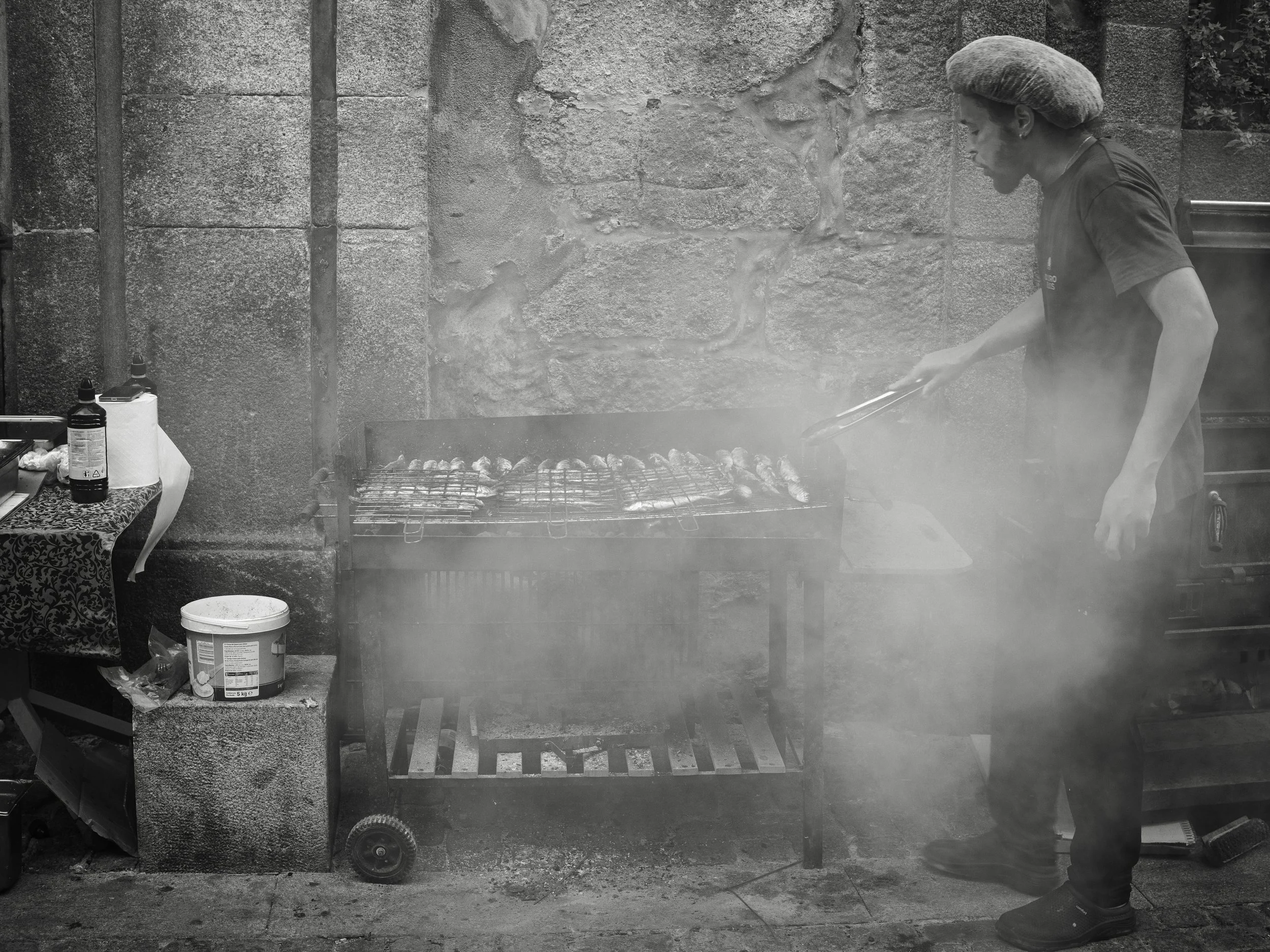 A person wearing a hat and black shirt is grilling food on a barbecue grill outdoors, surrounded by smoke. There is a stone wall and a table with bottles and other items nearby.