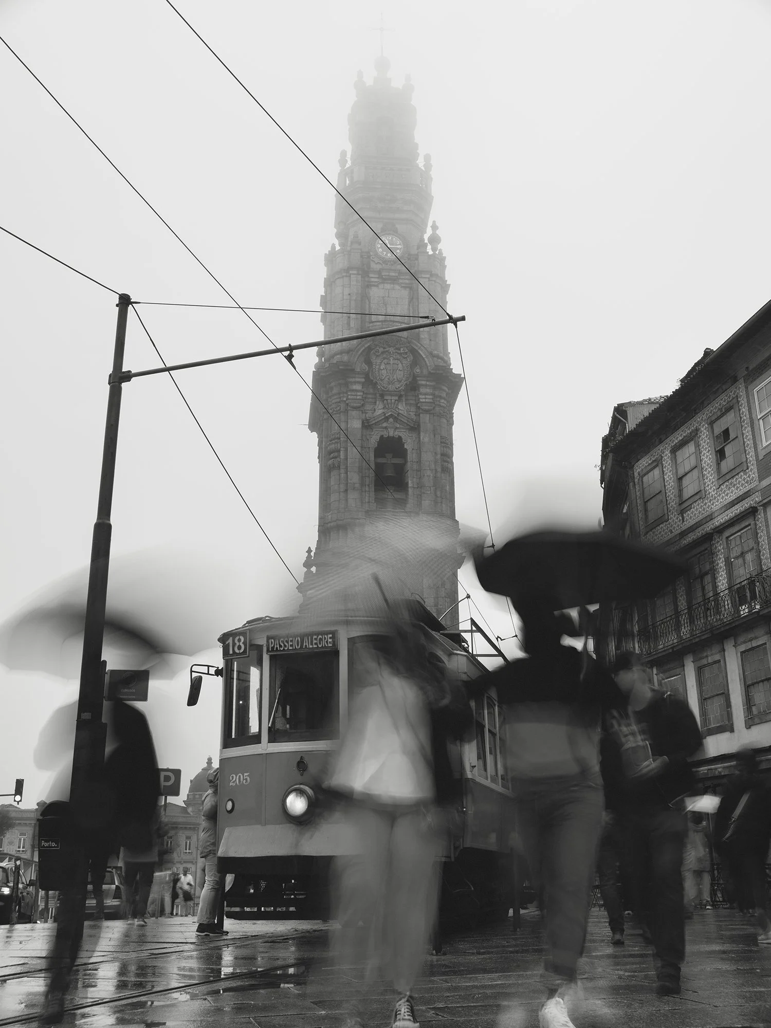 Black and white photo of a city street scene with pedestrians and a tram labeled 'Passeio Alegire.' A tall, foggy clock tower is visible in the background, along with historic buildings to the right.
