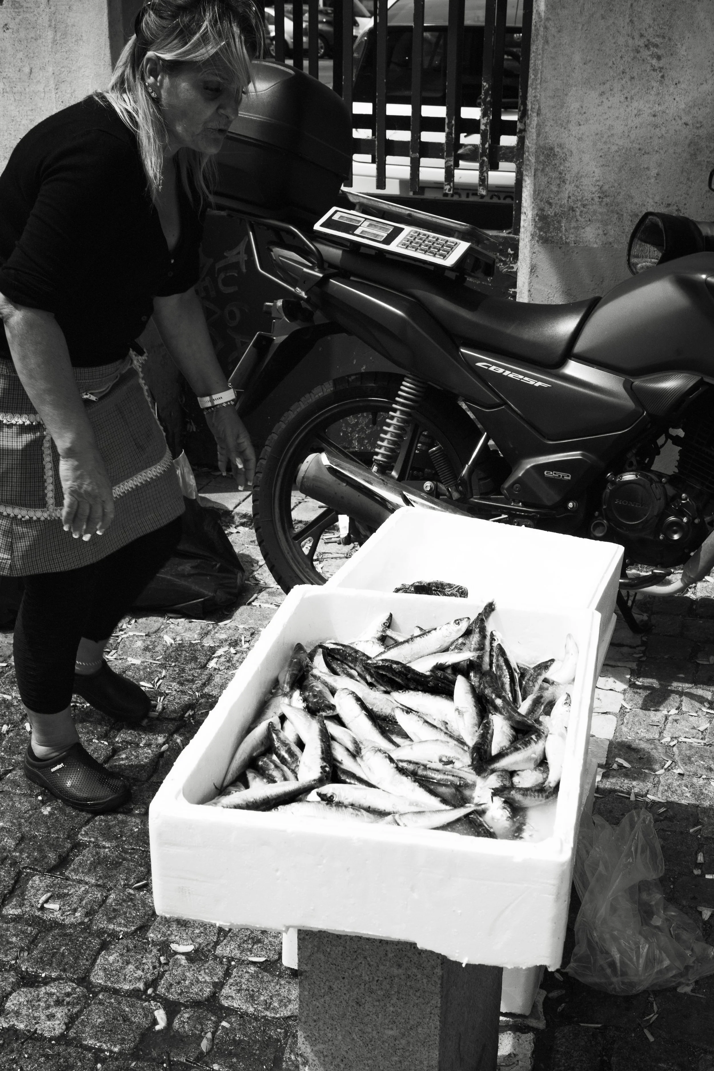 A woman with a checked apron looking at fish in a white styrofoam box outside near a motorcycle and a parking lot.