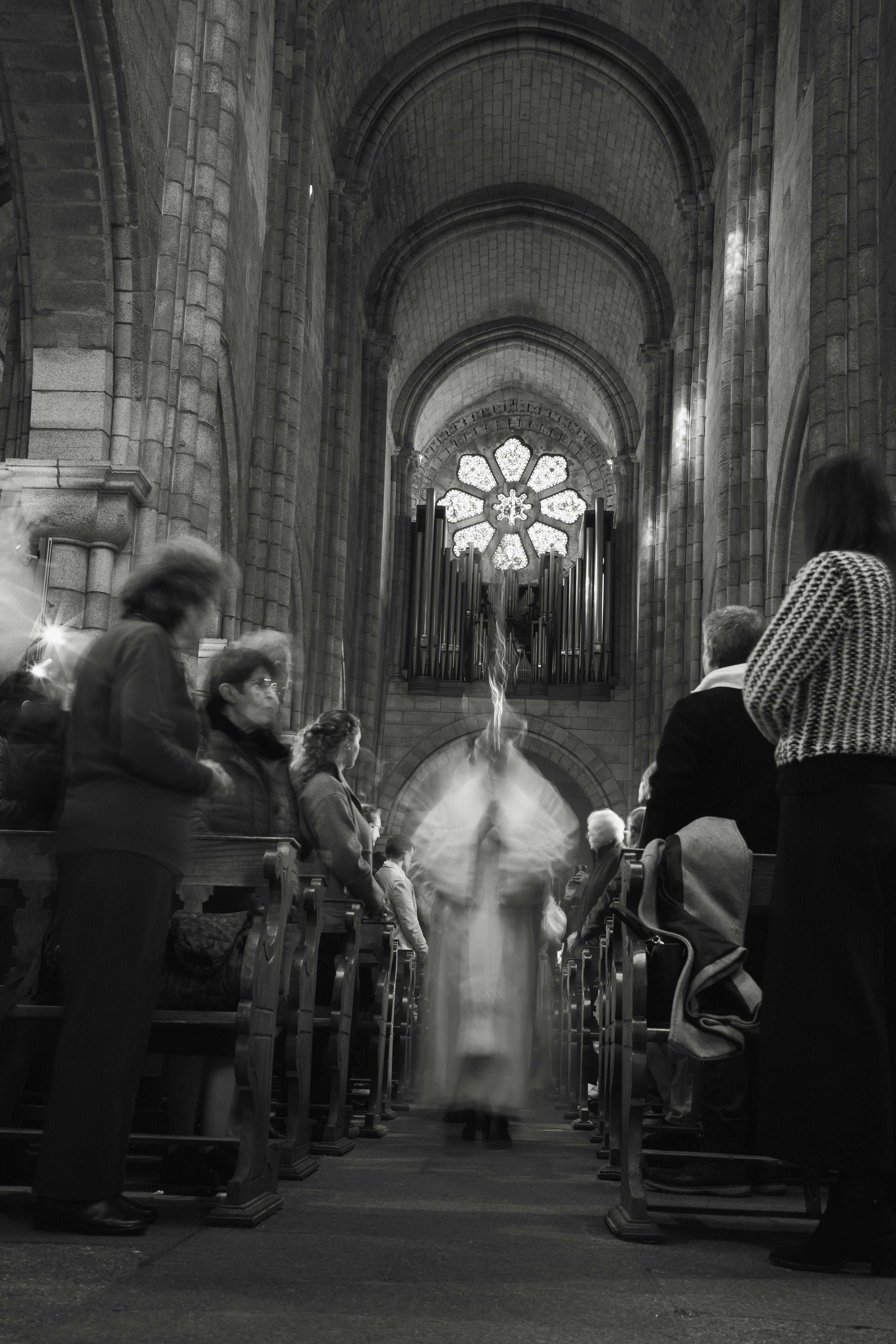 Inside a stone church with a large stained glass window and pipe organ, people standing and sitting in pews, blurred figure walking down the central aisle wearing religious robes.
