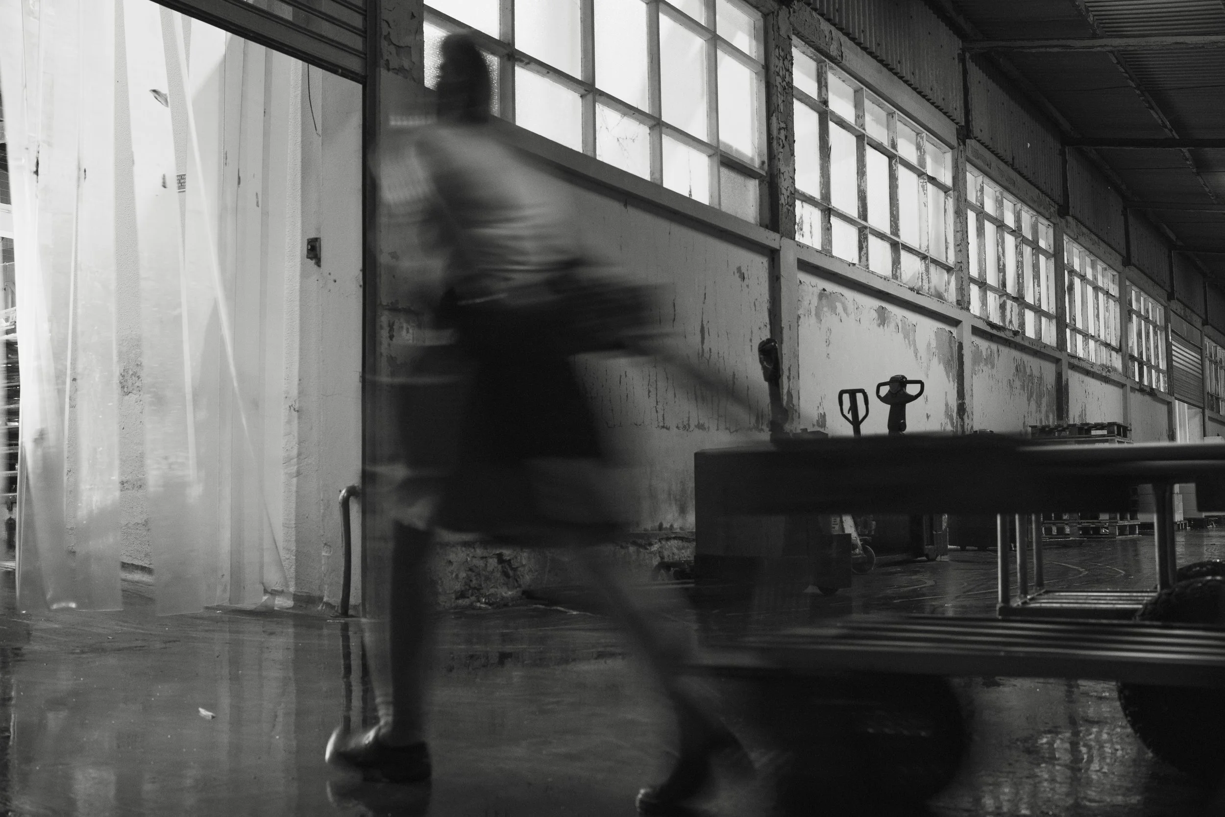 A blurry person walking past a large windowed industrial building interior, with a table and some tools visible.