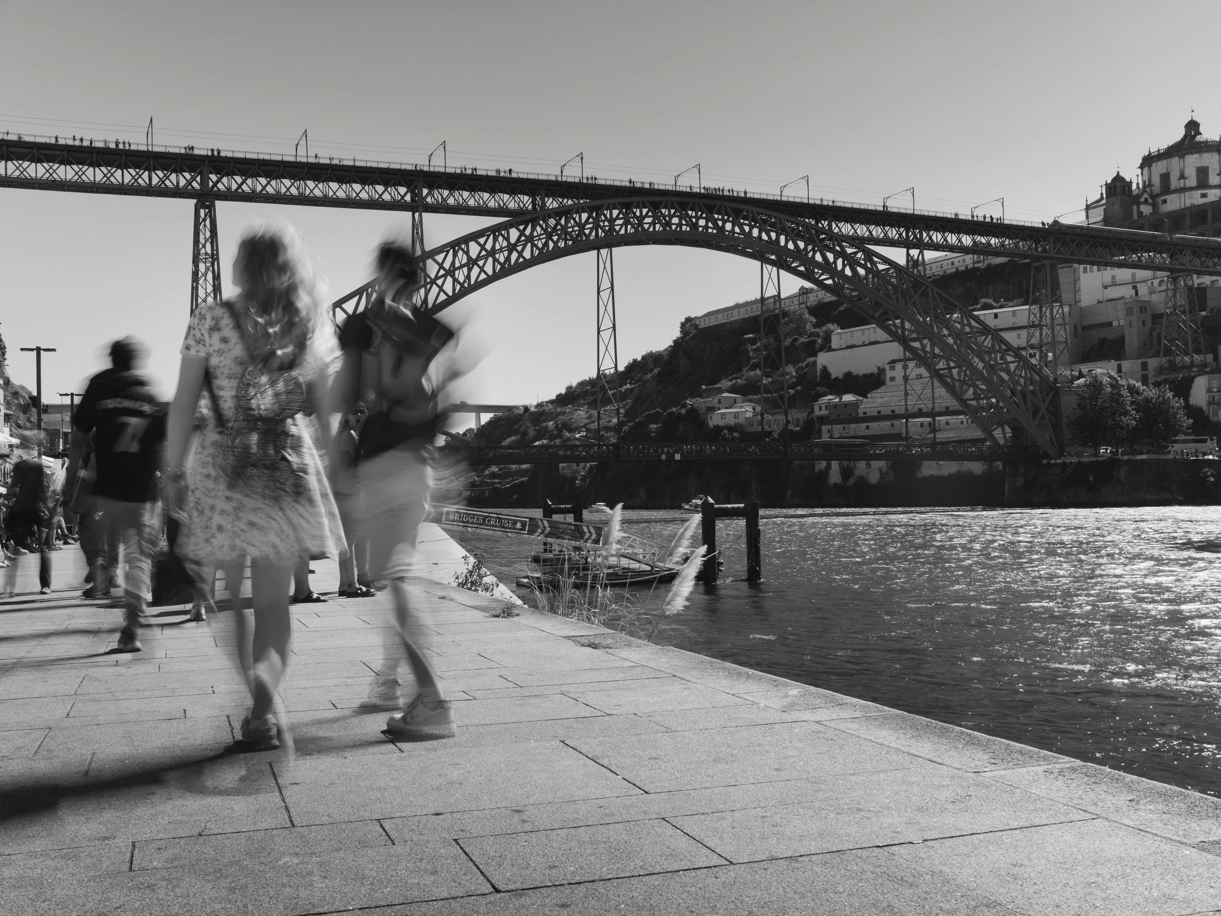 People walking along a riverfront path near a large bridge with an arch and a hillside with buildings in the background. The photo is in black and white.