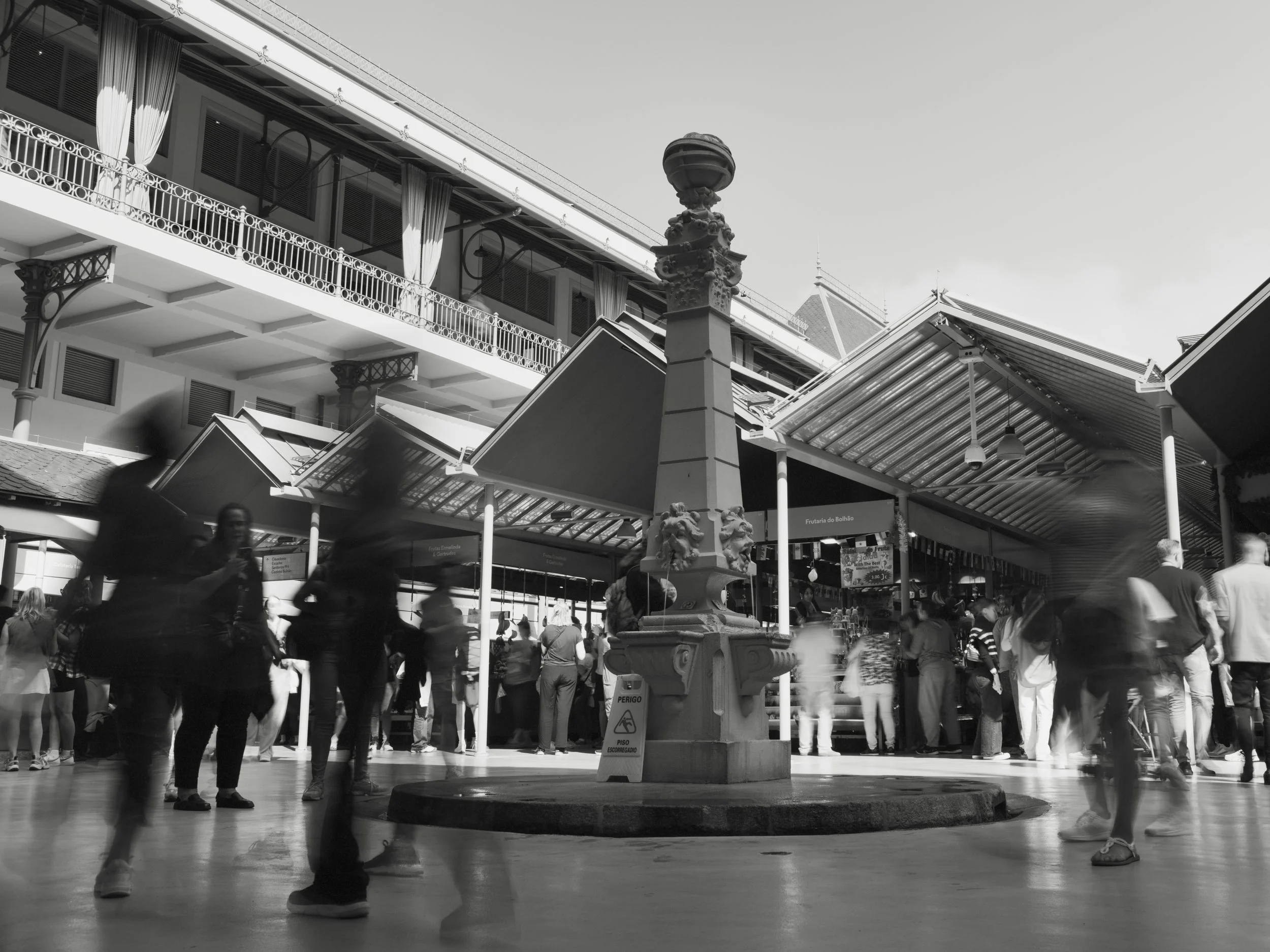 A busy indoor market area with a central fountain and numerous people walking or standing around, some blurred from motion. The market is housed in a building with balconies and decorative architecture.