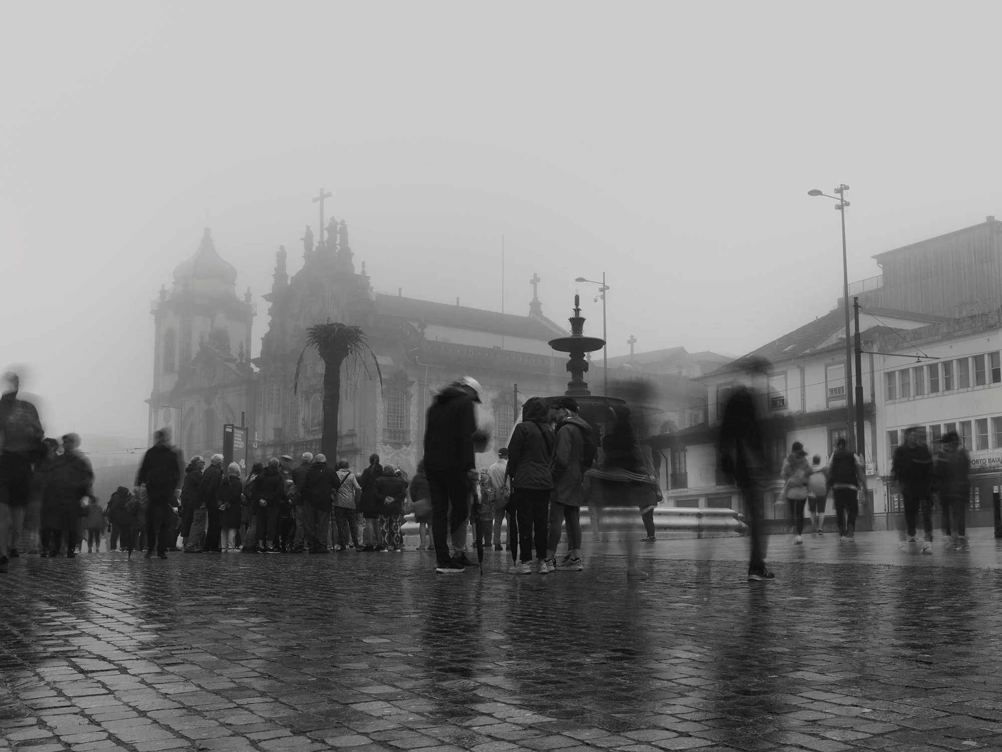 A black and white photo of a foggy city square with a crowd of people and historic buildings in the background, including a church with domes and crosses.