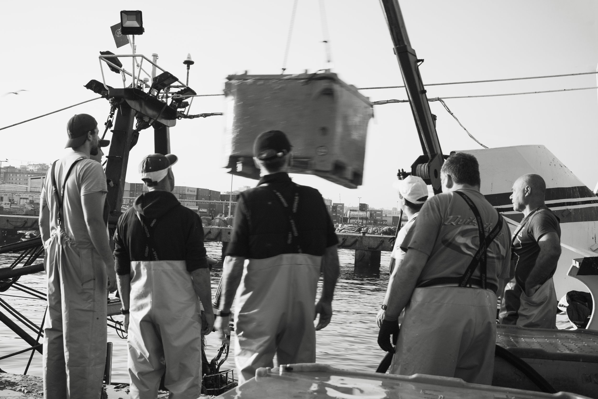 Group of people working with a crane to lift a large container or box near a body of water, with ships and industrial buildings in the background.