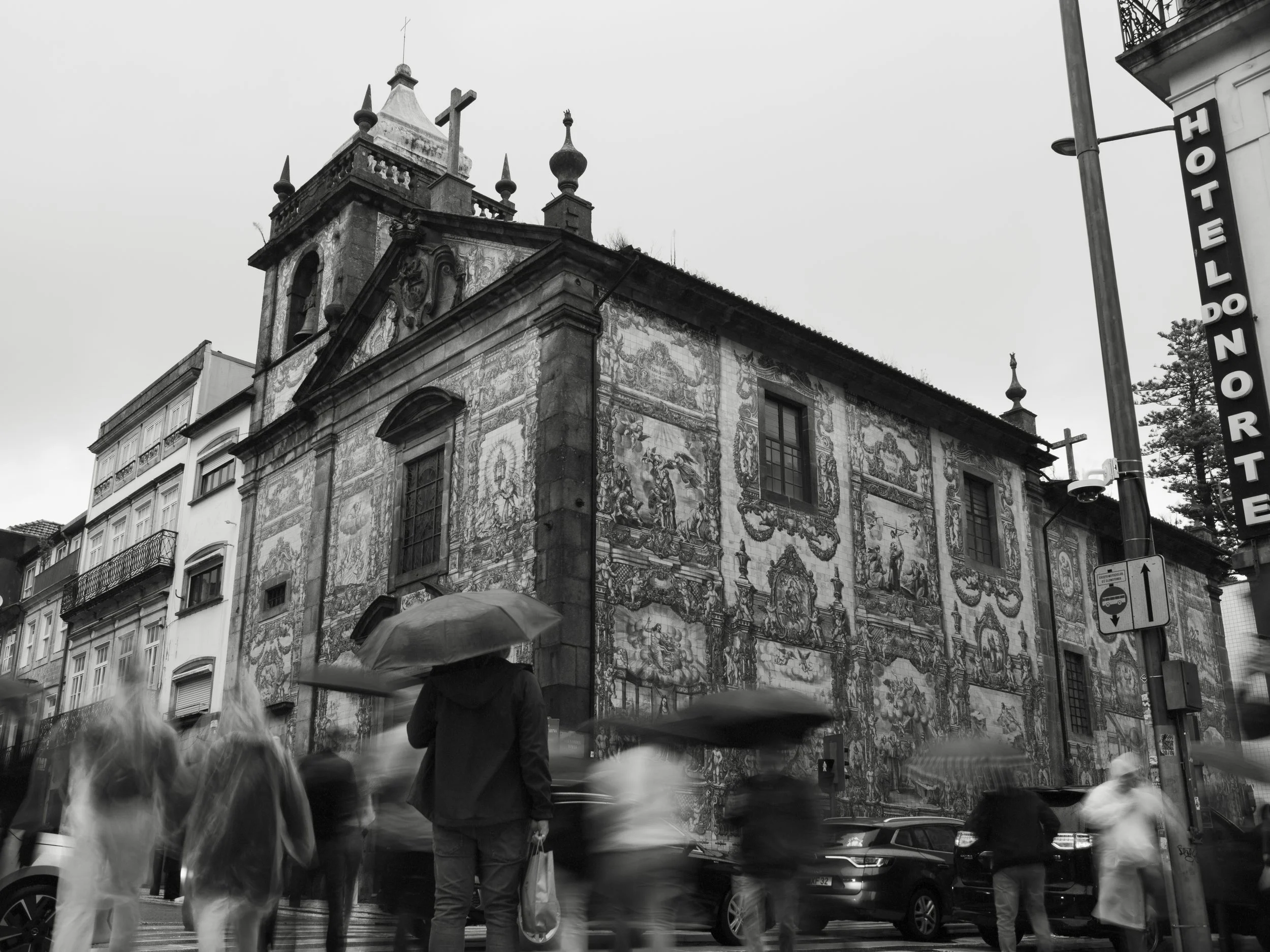 Street scene in front of a historic church with detailed facade artwork, people with umbrellas crossing the street, and cars parked along the curb.