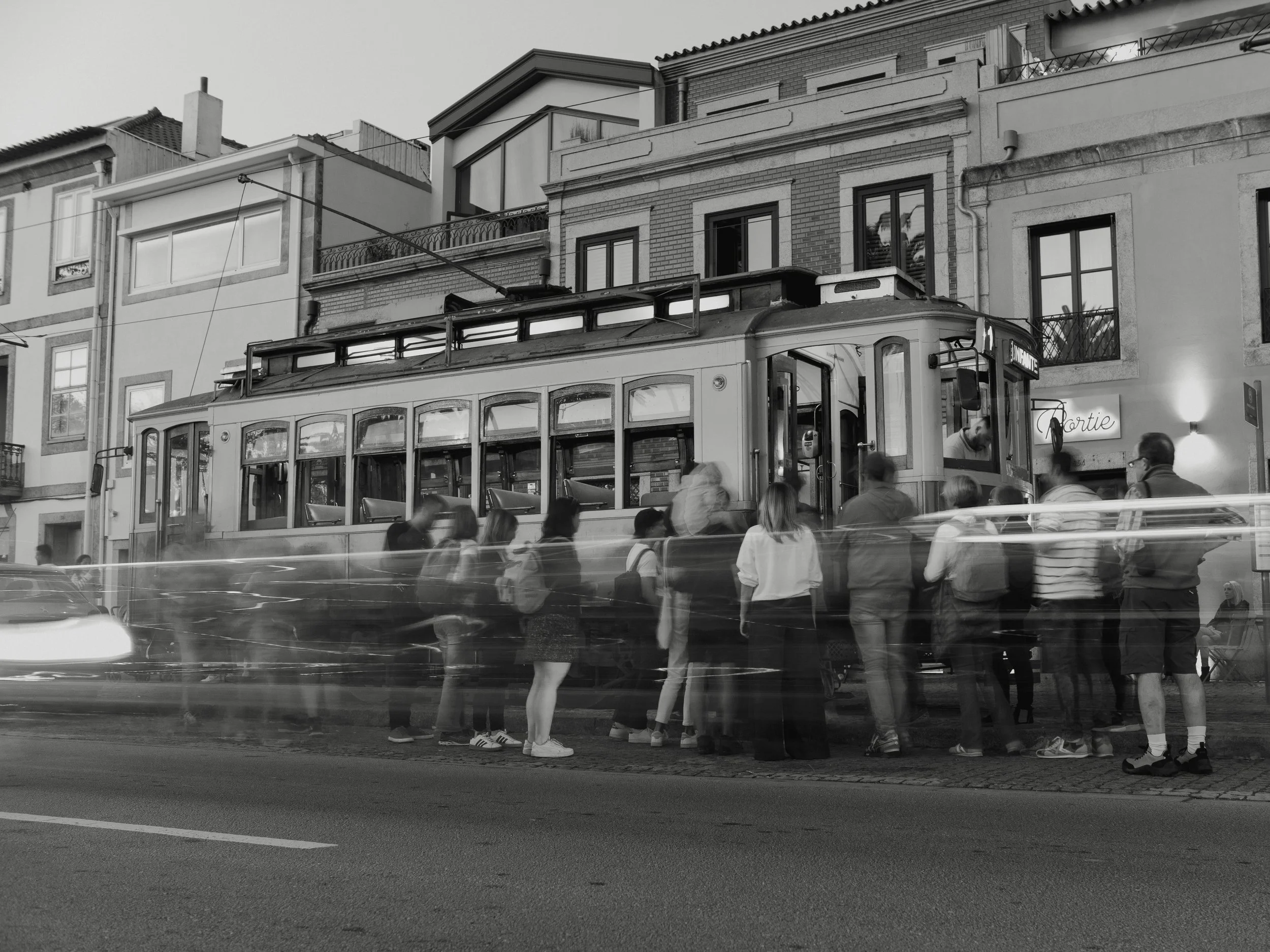 A city street with a vintage trolley car stopped at a stop. People are waiting in line to board or ride, some standing on the sidewalk and some climbing into the trolley. There are residential buildings in the background and motion blur of passing ve