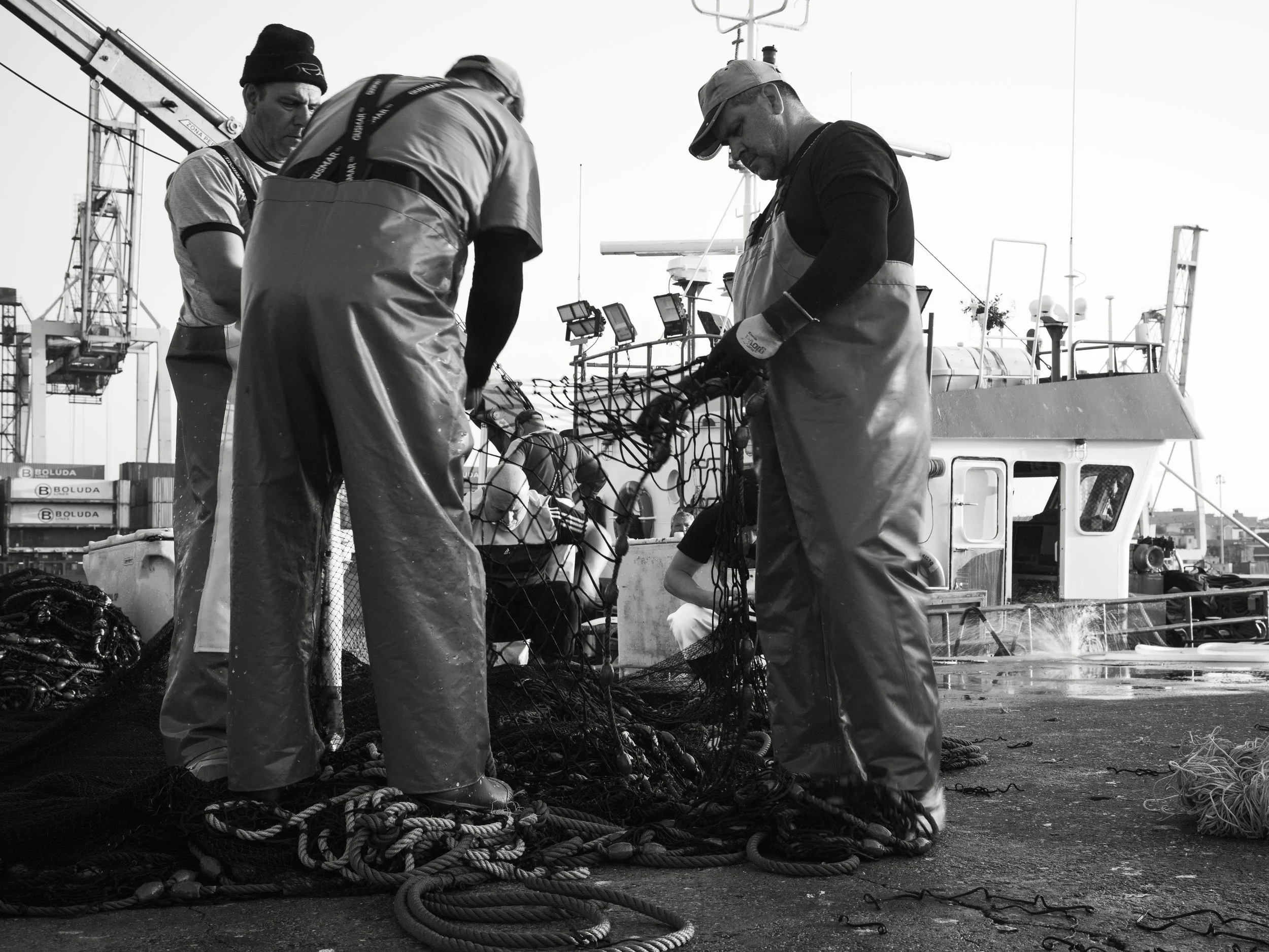 Three fishermen in waterproof overalls working together to untangle fishing nets on a dock next to a boat.