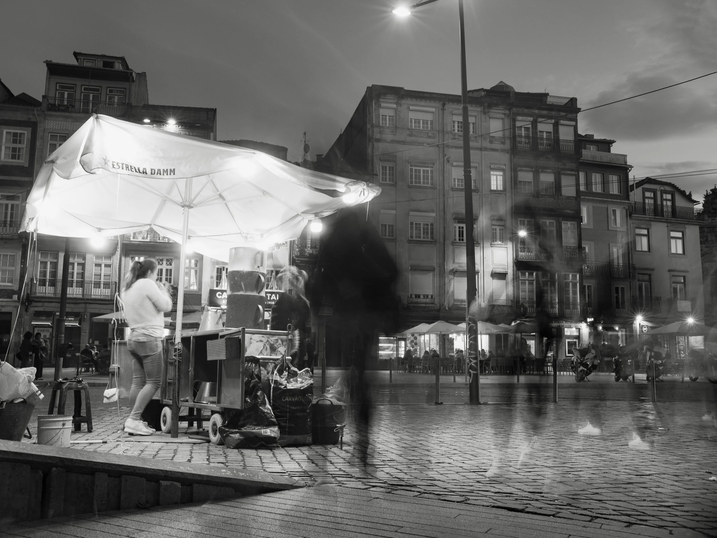 Nighttime street scene with a small food stand under a large umbrella, people walking by, and a row of multi-story buildings in the background.