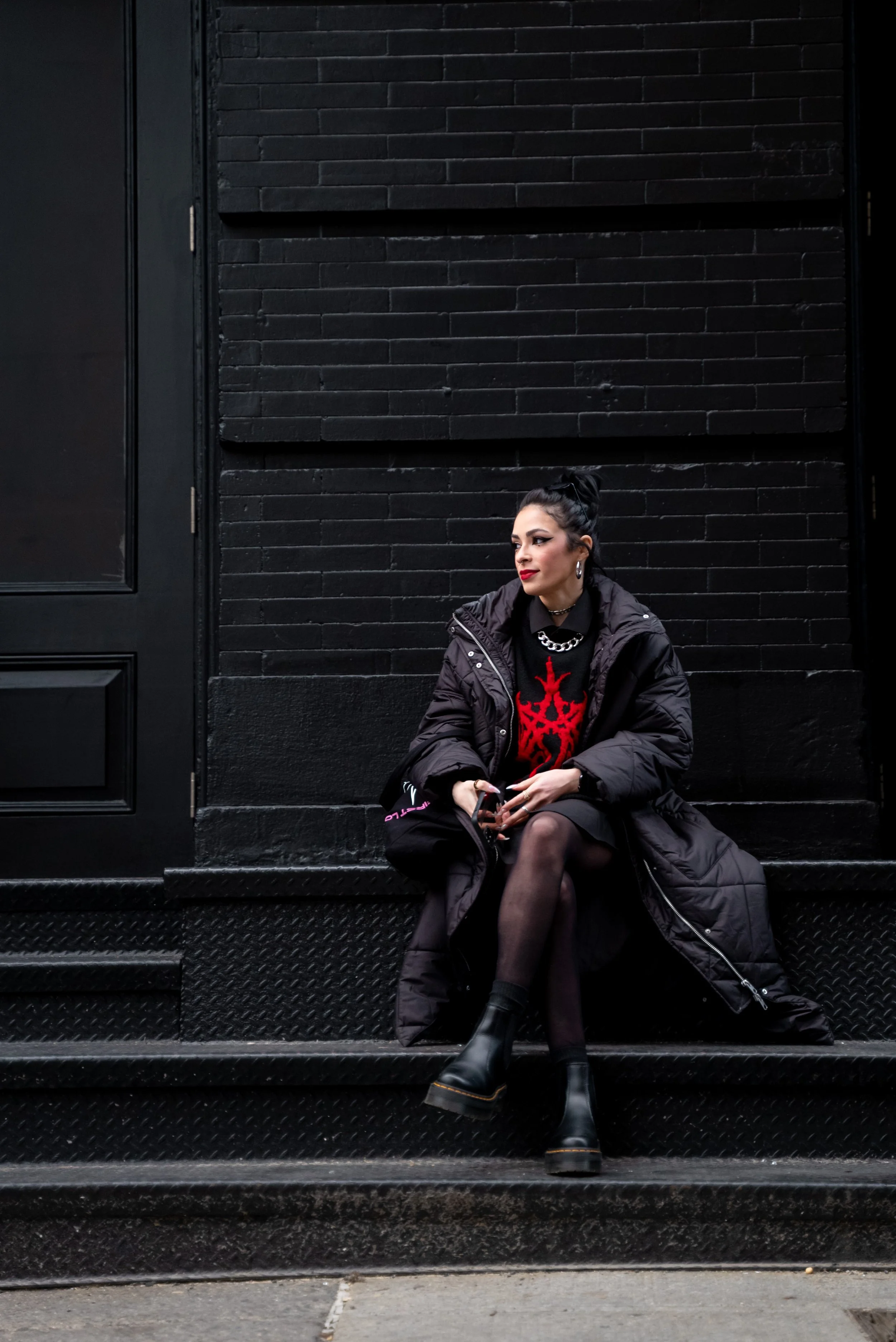 A woman sitting on black stairs outside a black building, wearing a black coat, a black dress with red design, black boots, and jewelry, with dark hair tied back.