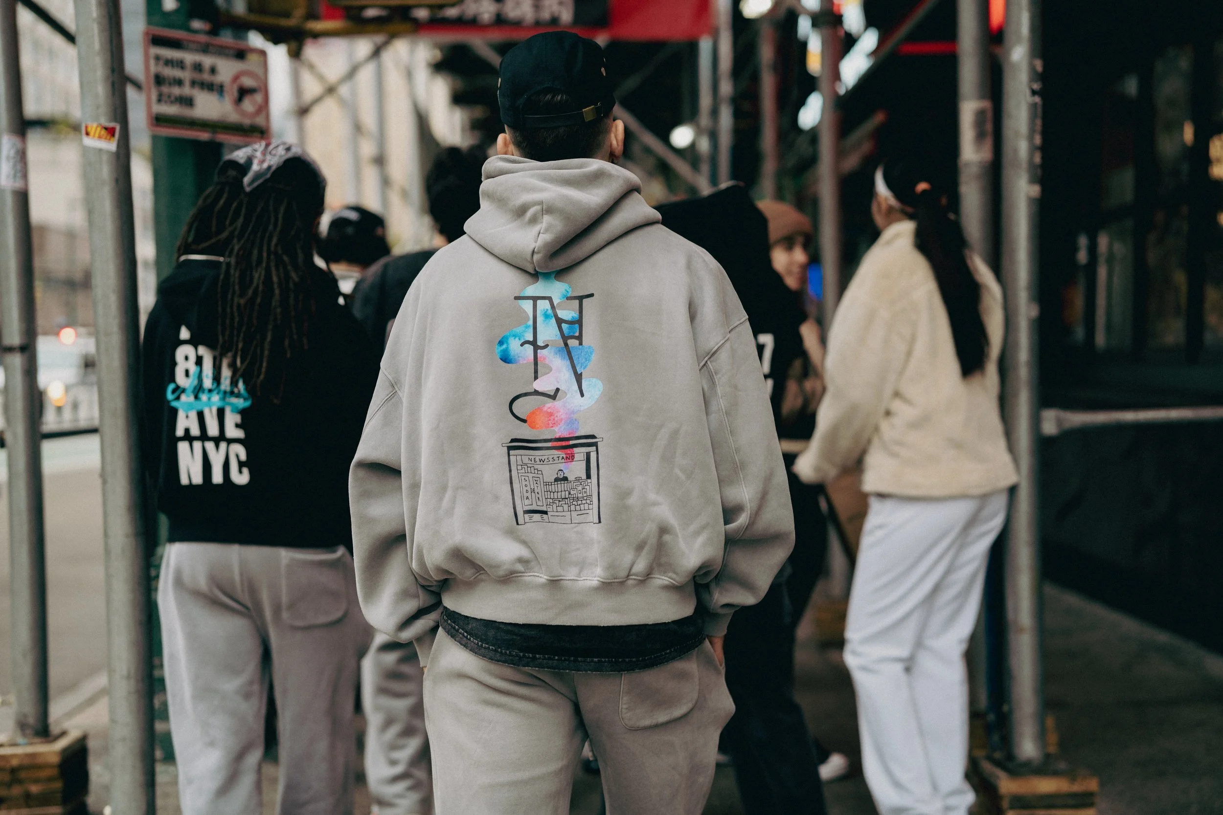 A group of people standing on a city sidewalk, with one person wearing a gray hoodie featuring a graphic of a newspaper and colorful smoke artwork on the back. Others are dressed casually, and some are wearing face masks.