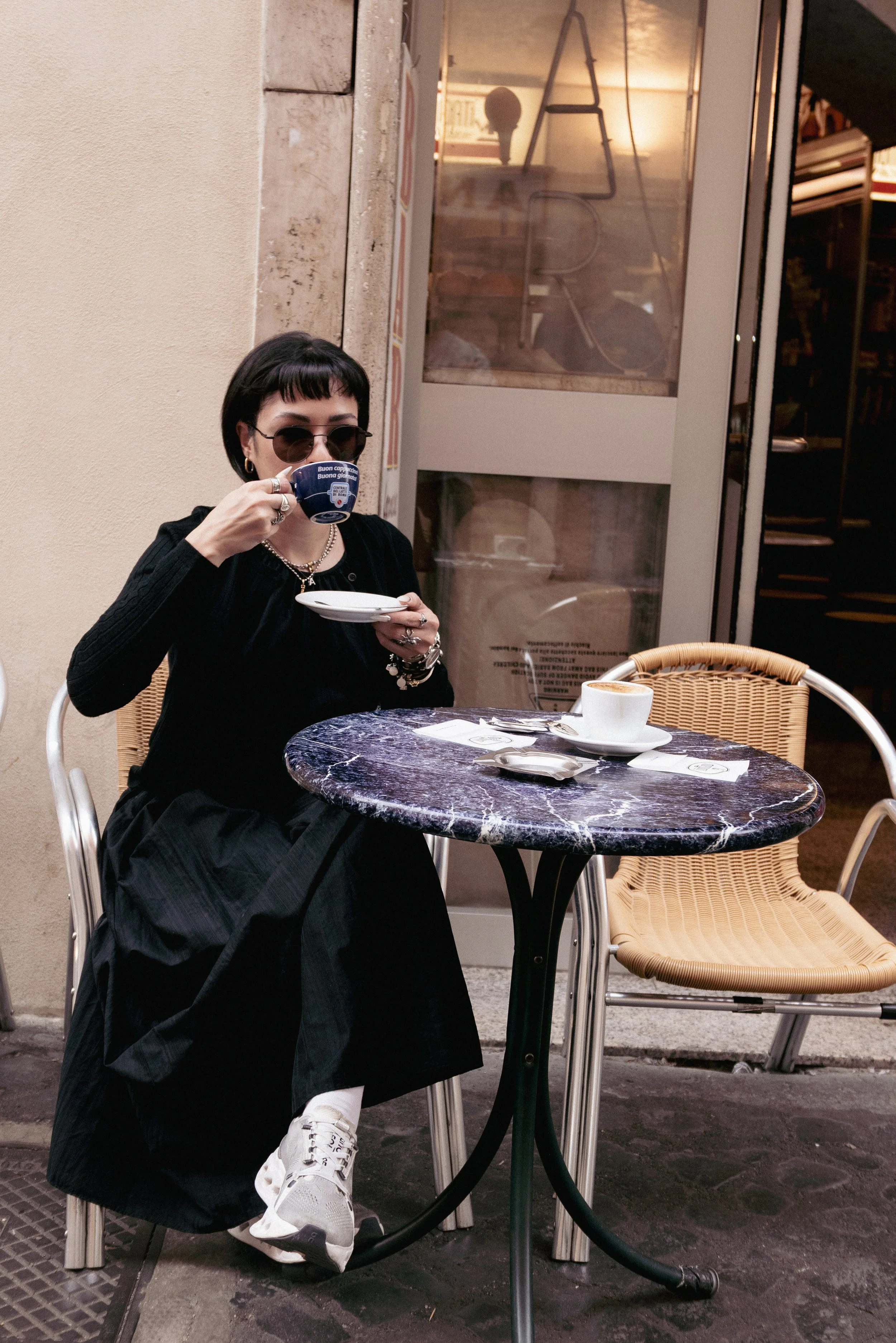A woman sitting at an outdoor cafe table, drinking from a coffee cup, wearing dark sunglasses, a black outfit, with a marble top table, and a small plate and spoon on the table.