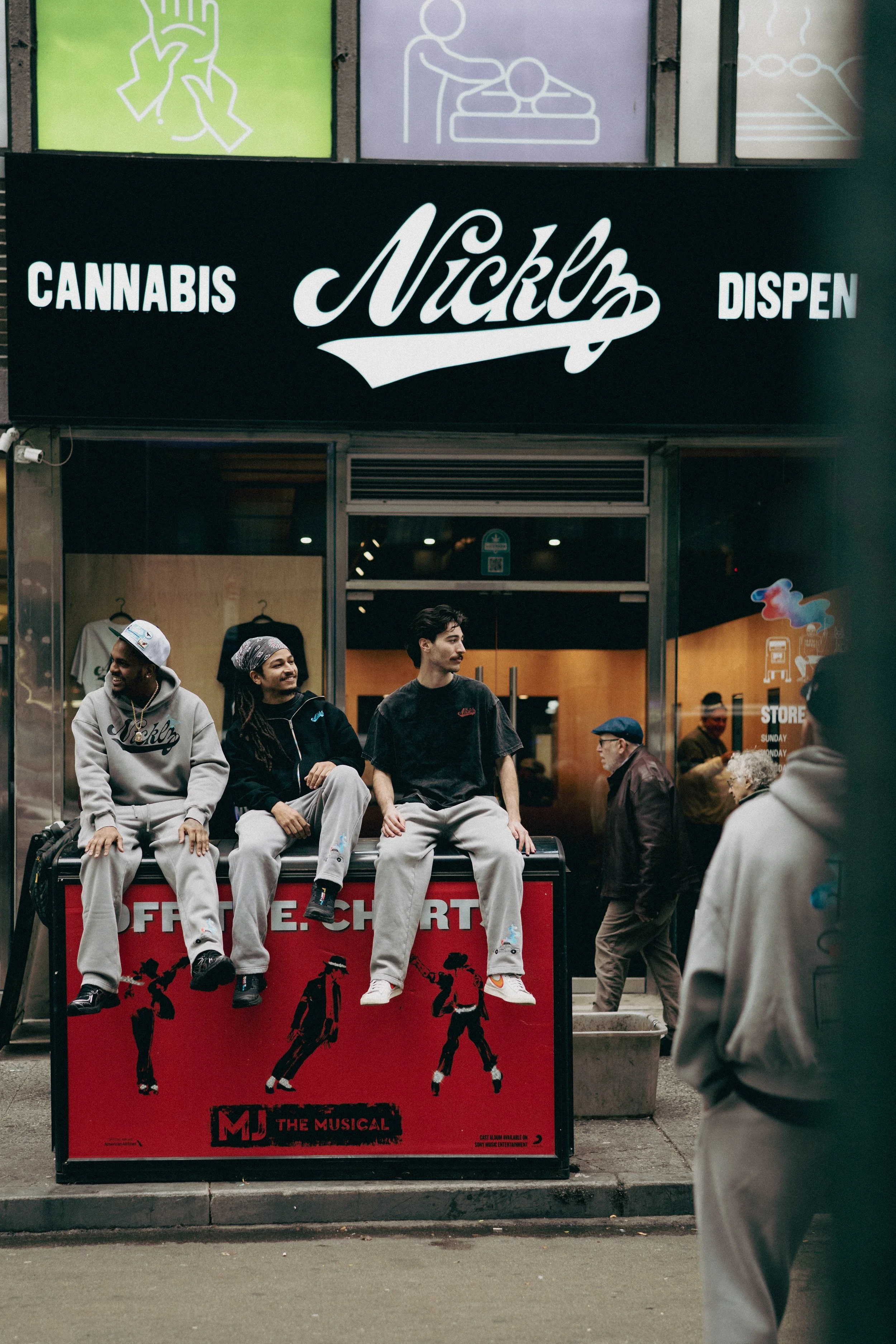 Three young men sitting on a red advertising board outside a cannabis dispensary named 'Nickel'. The men are dressed in casual streetwear, and there are people walking by in the background.