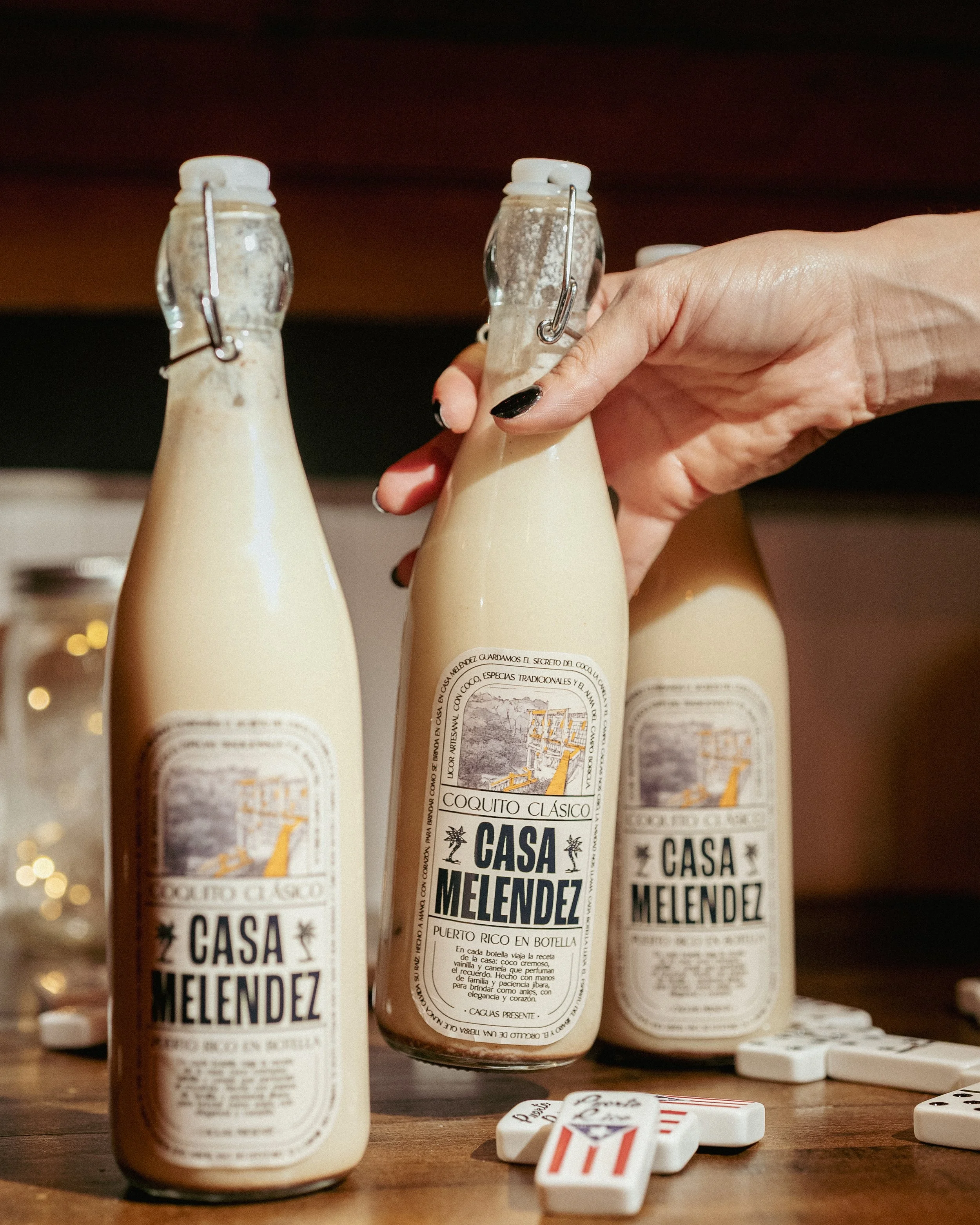 Hand holding a bottle of Casa Melendez Coquito Classic, with two other bottles of the same beverage on a wooden surface surrounded by domino pieces.