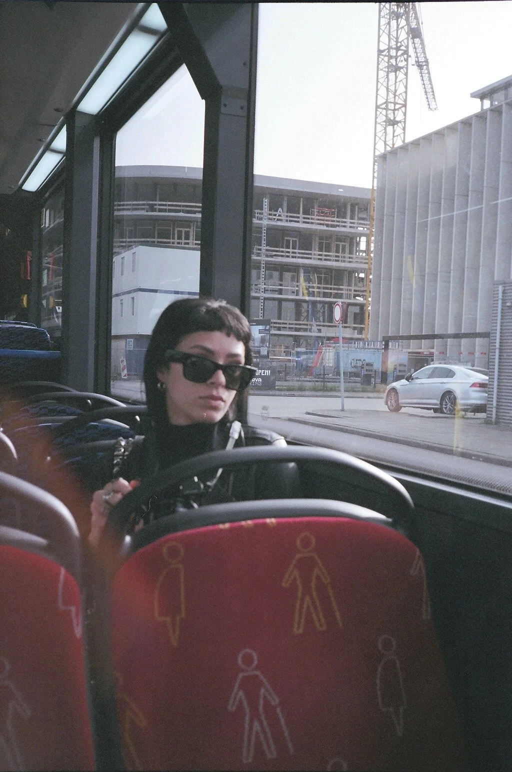 A woman with short dark hair and sunglasses sitting on a bus, holding onto a strap. The bus has colorful seats with walking person symbols, and the background outside the window shows a construction site with buildings, a crane, and parked cars.