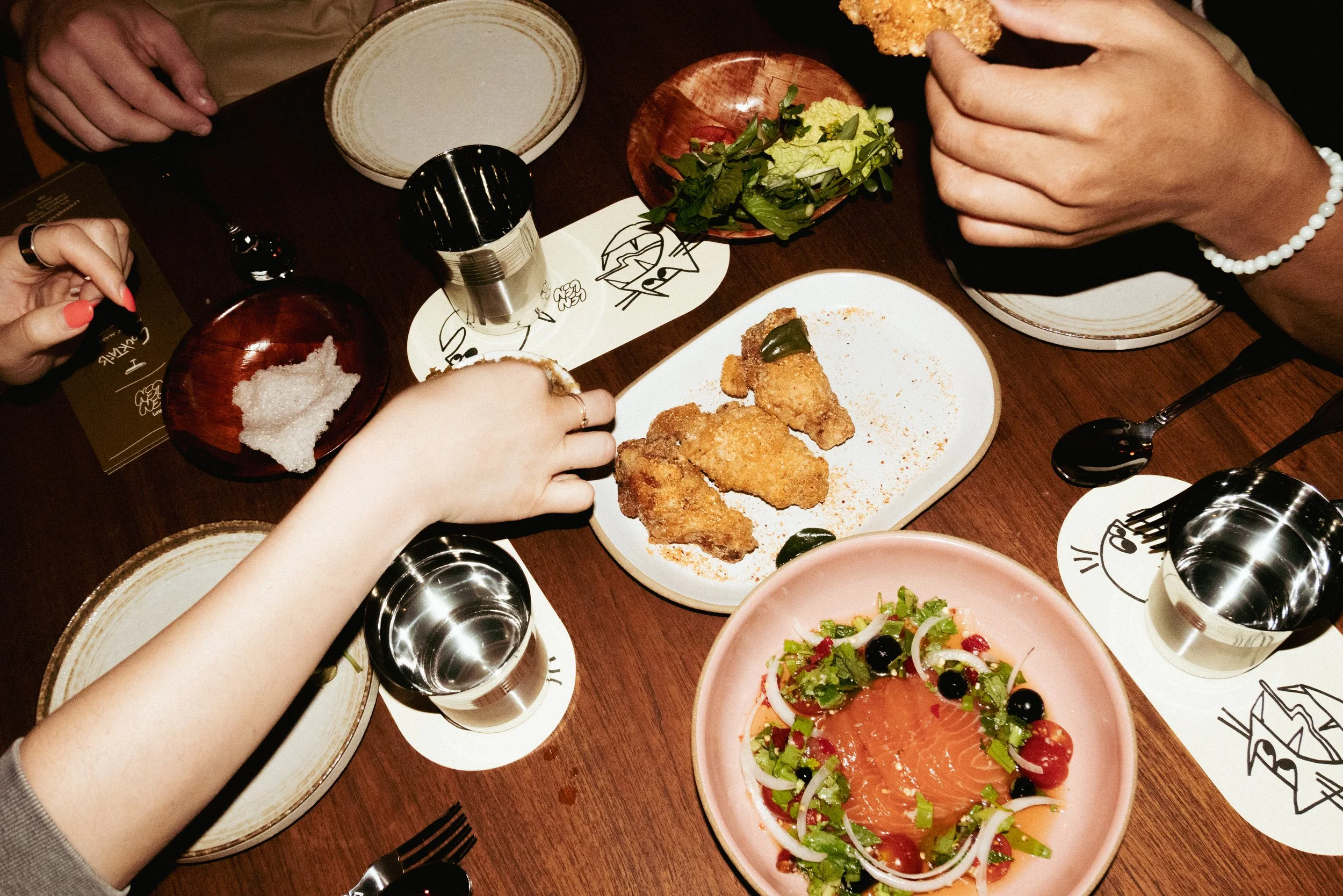 People sharing a meal at a dinner table with fried chicken, a salad with salmon, salt, salt shakers, and empty plates.