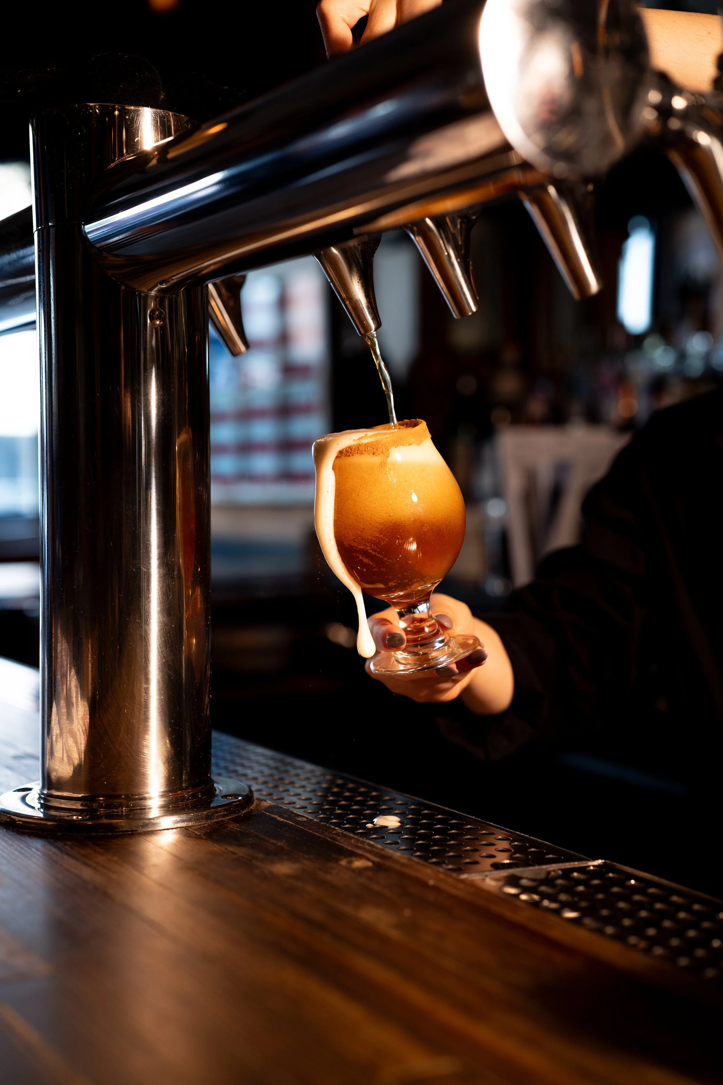 A person holding a glass of beer under a tap in a bar, with foam dripping down the side of the glass.