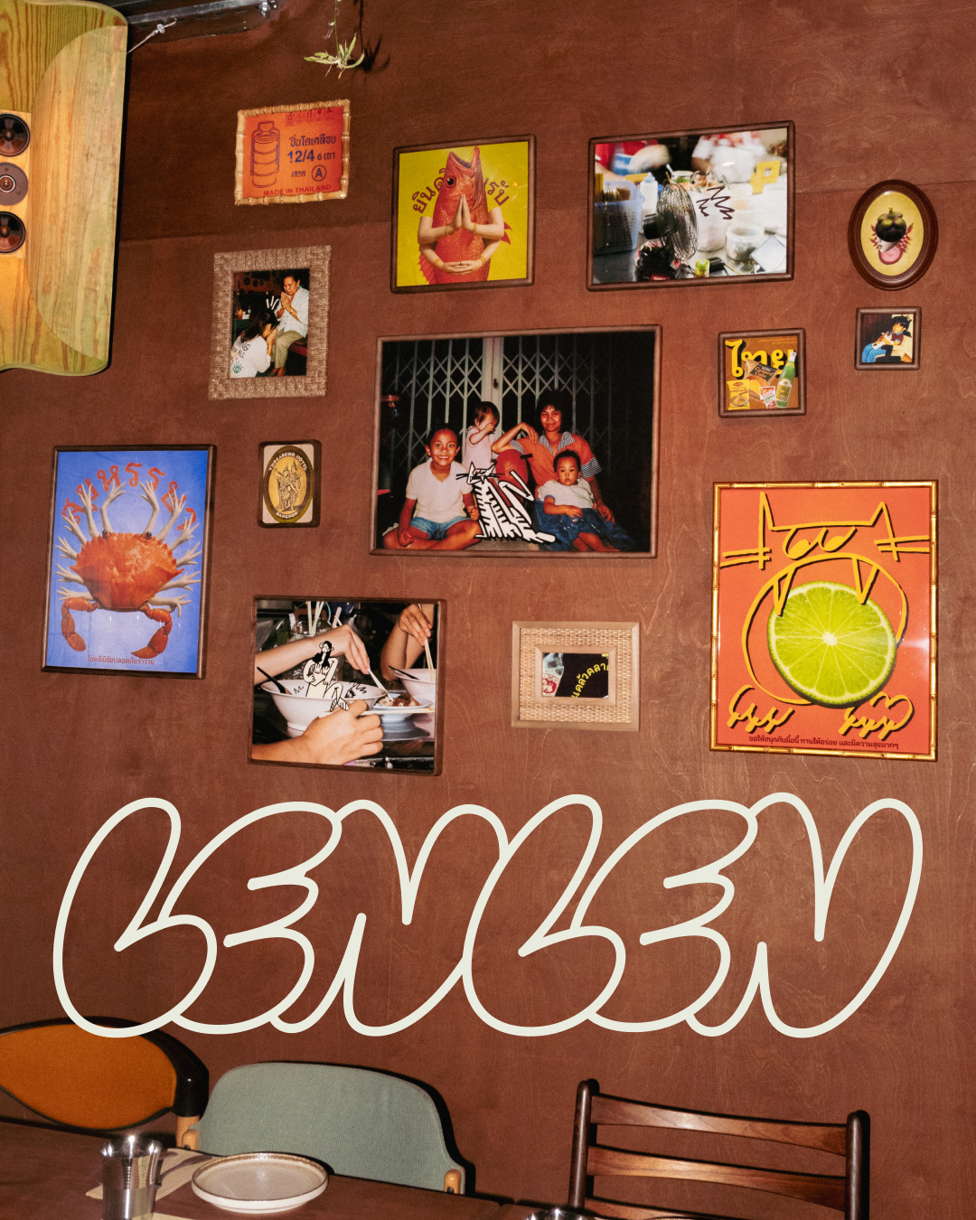 Wall decorated with various framed photographs and posters, including images of people, food, and Thai-themed artwork. Part of a dining table and chairs are visible at the bottom, with some utensils and a plate on the table.