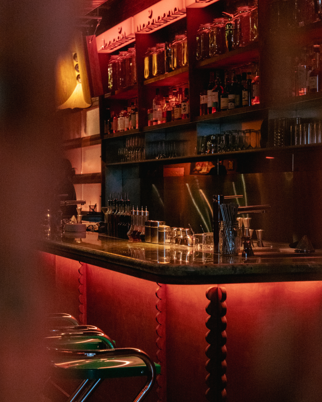 A dimly lit bar with shelves of liquor bottles, glassware, and bar tools, illuminated by warm red lighting, with bar stools in the foreground.