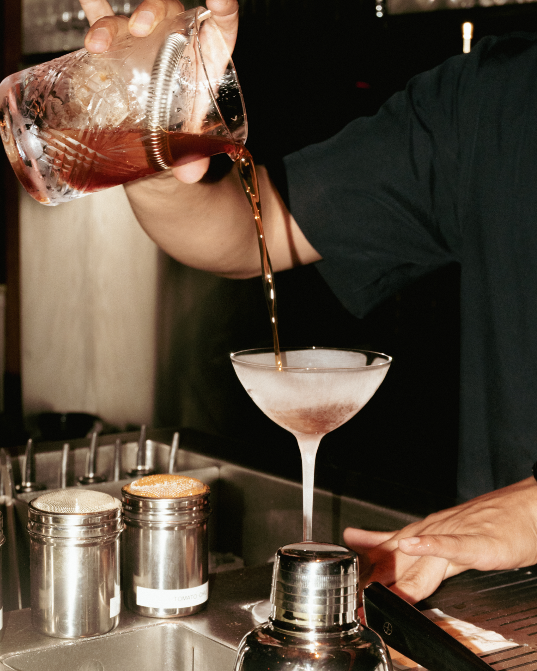 A bartender pours red mixed drink from a measuring shaker into a martini glass at a bar, with various bar tools and containers visible on the counter.