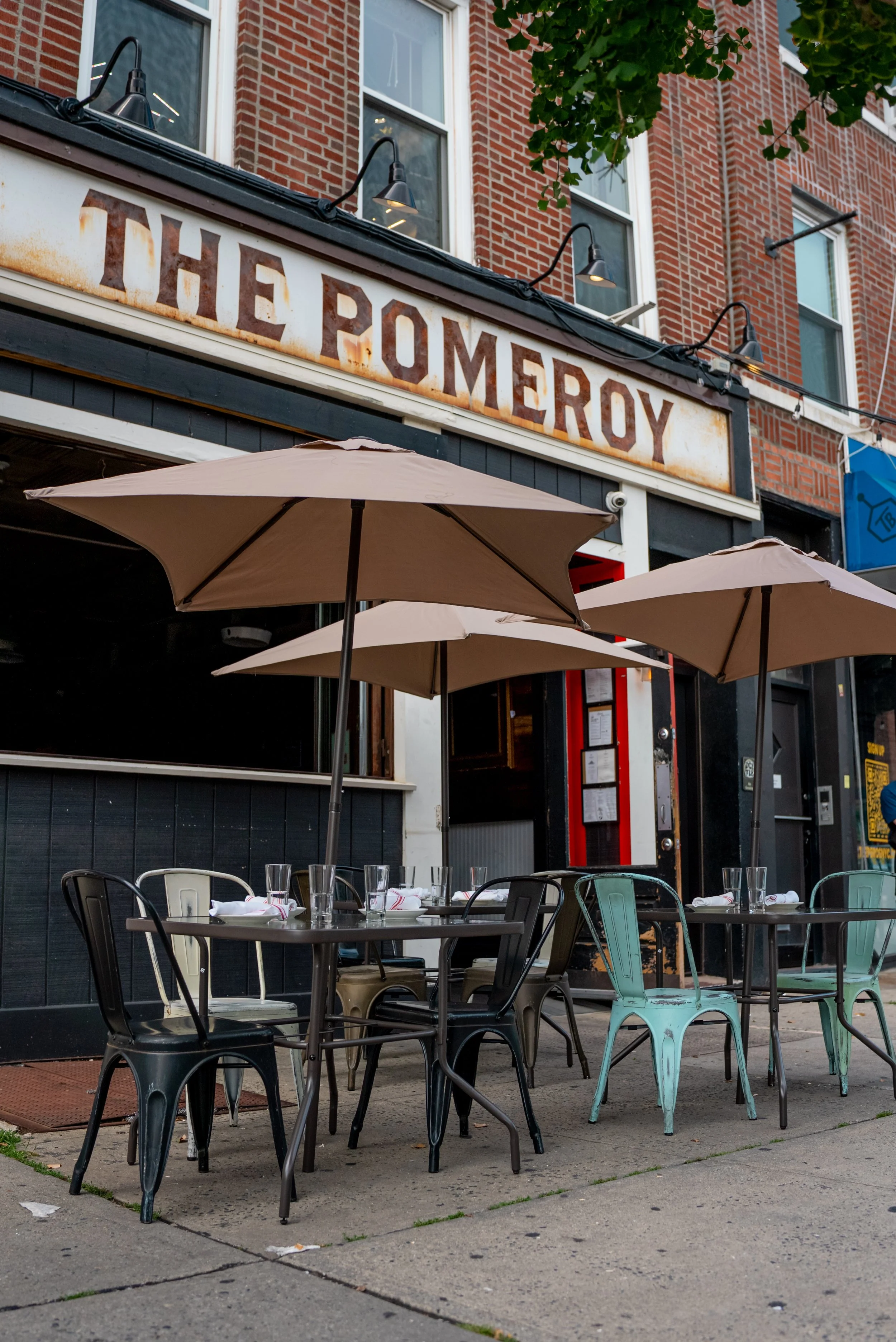 Empty outdoor seating area with tables, chairs, and umbrellas outside a restaurant called The Romero with a brick building and tree in the background.
