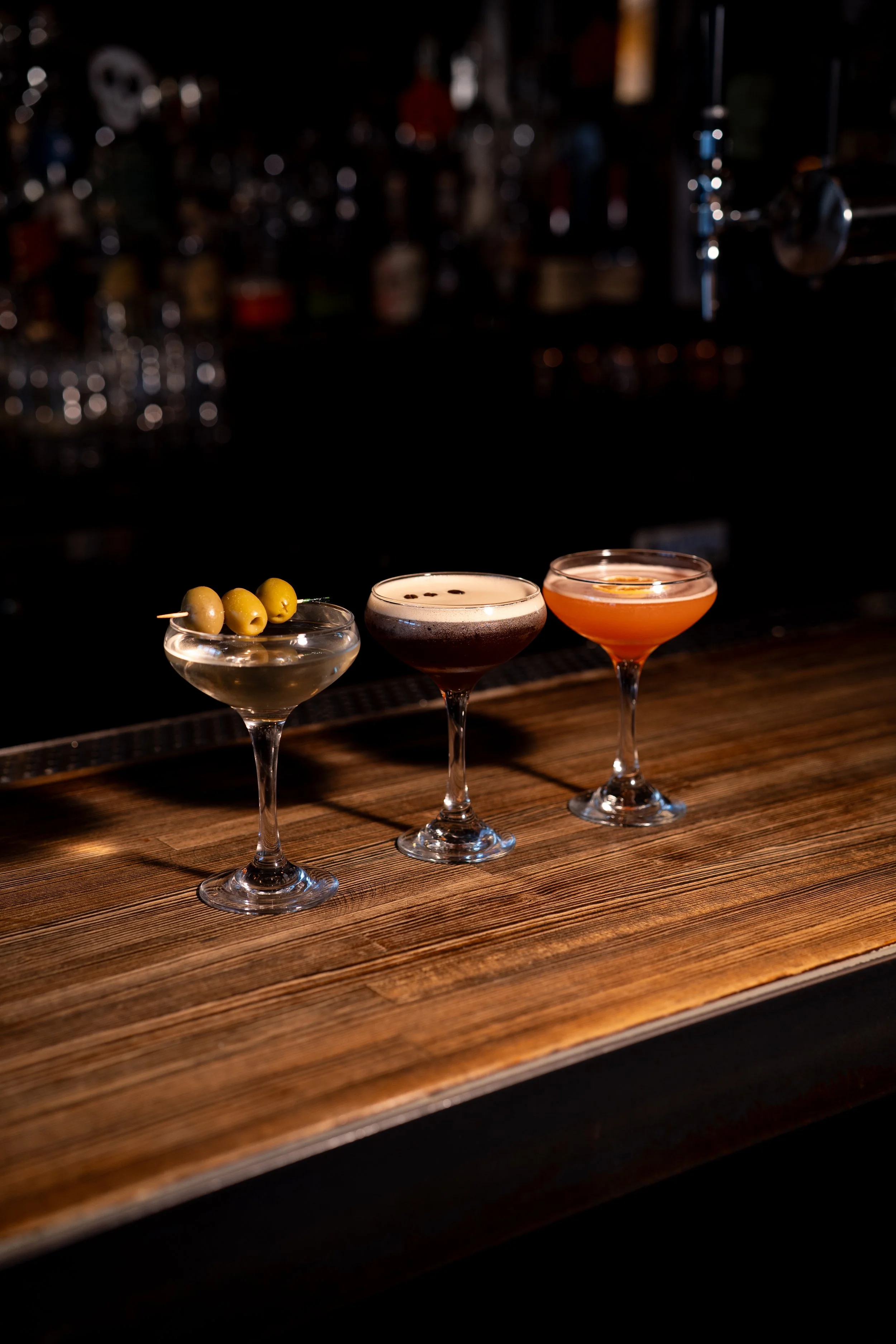 Three cocktails on a wooden bar counter with a dark, blurry bar background.
