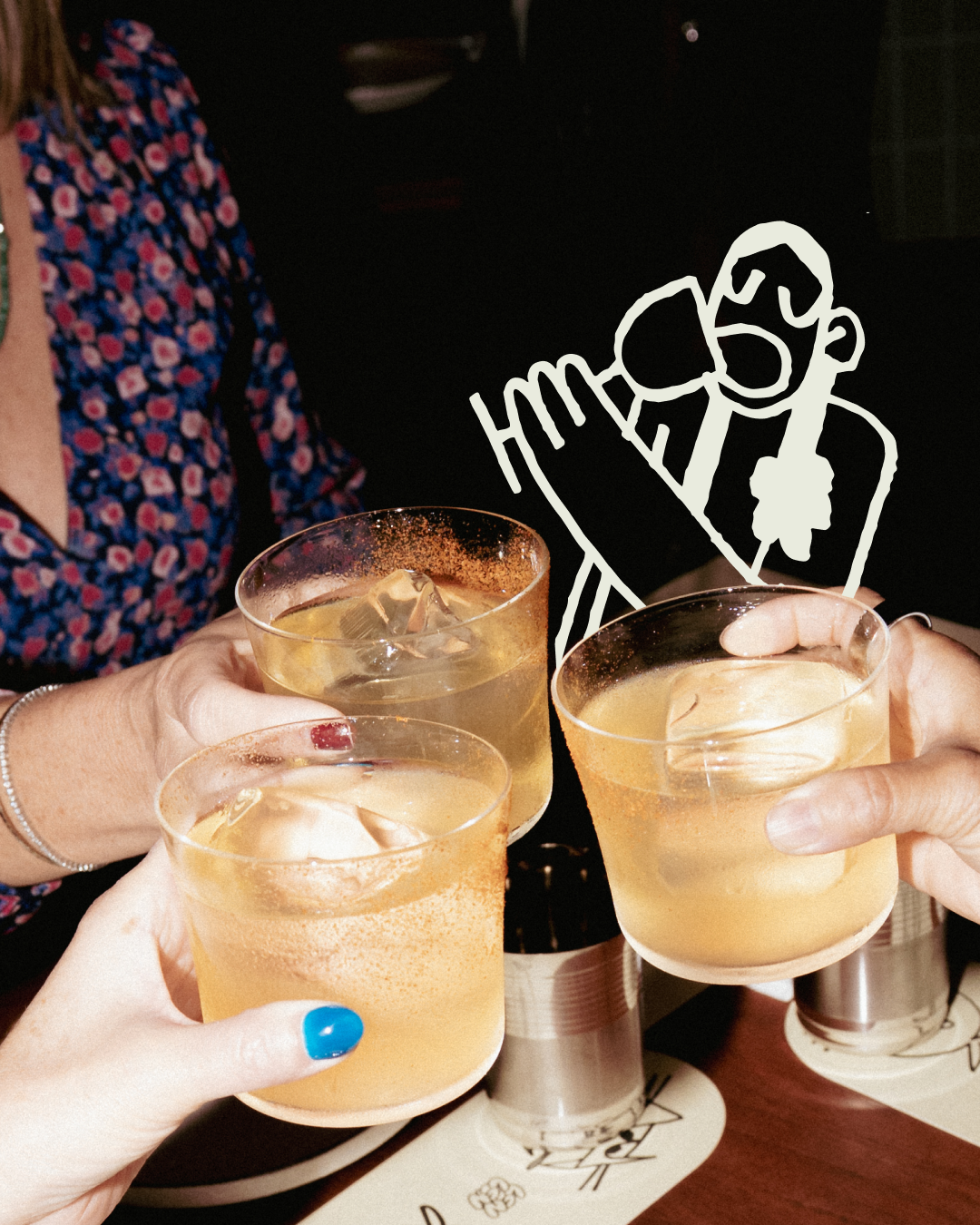 Three people holding glasses of a light-colored cocktail with ice, raising them for a toast. One person has blue nail polish, another has red, and the third person's hand is visible. In the background, there is a black and white outline drawing of a 