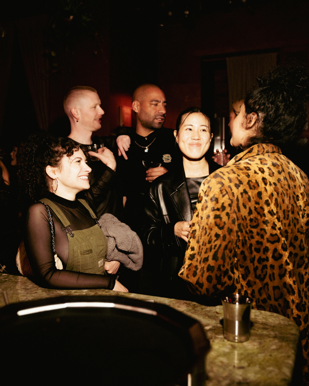 Group of six diverse friends socializing at a bar or nightclub, smiling and engaged in conversation, with some holding drinks, in a dimly lit, lively setting.