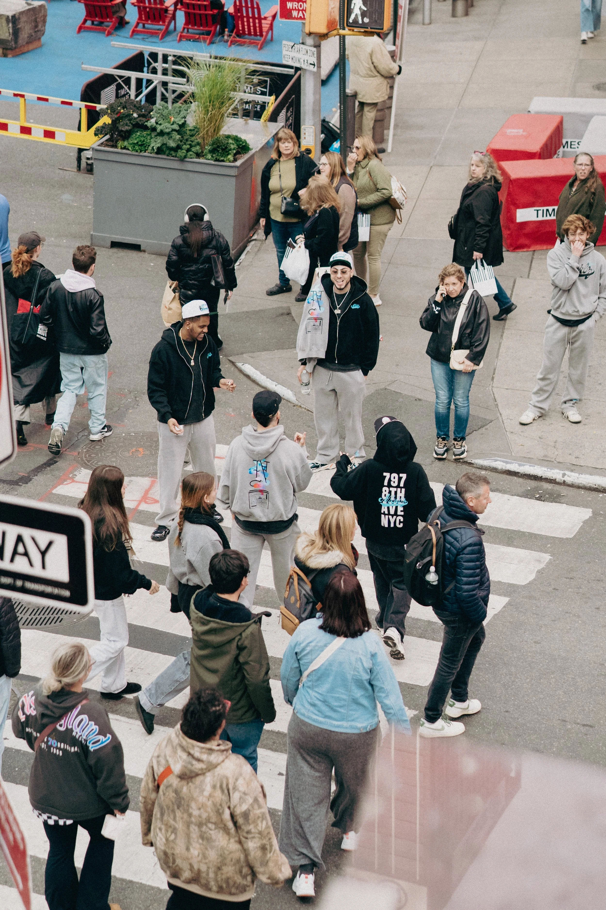 People crossing a city street at a crosswalk, some talking on phones, others walking in groups or alone, with storefronts and street signs visible.