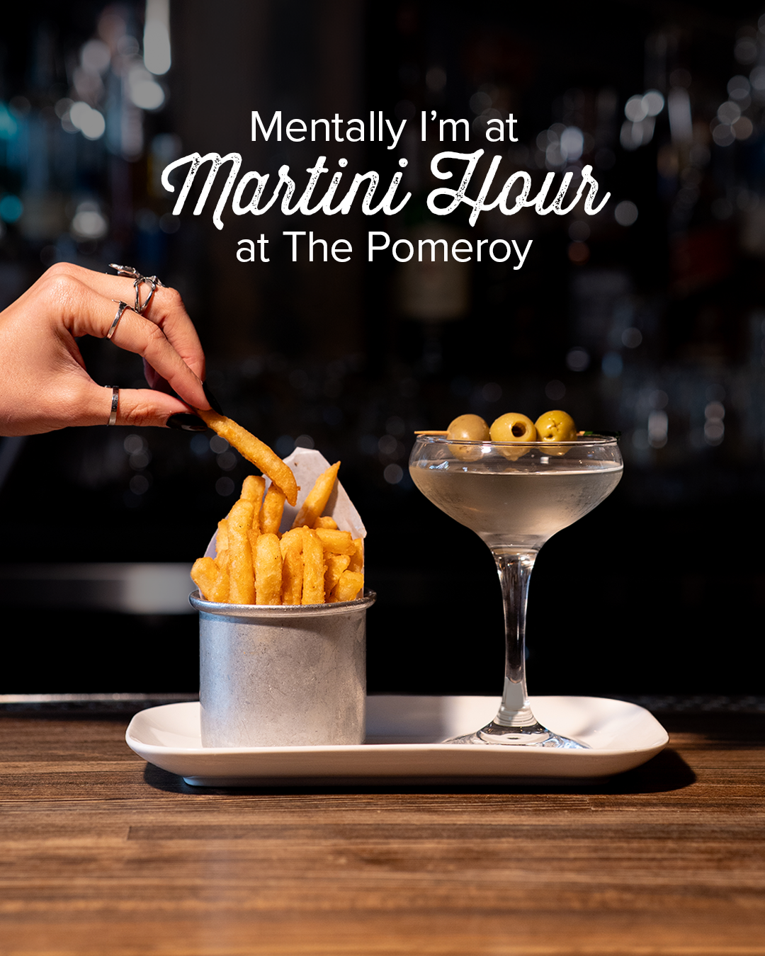 A hand reaching into a small metal container of French fries on a white serving tray, with a cocktail garnished with green olives next to it, set on a wooden bar counter background with an overlay text that reads 'Mentally I'm at Martini Hour at The Pomeroy.'